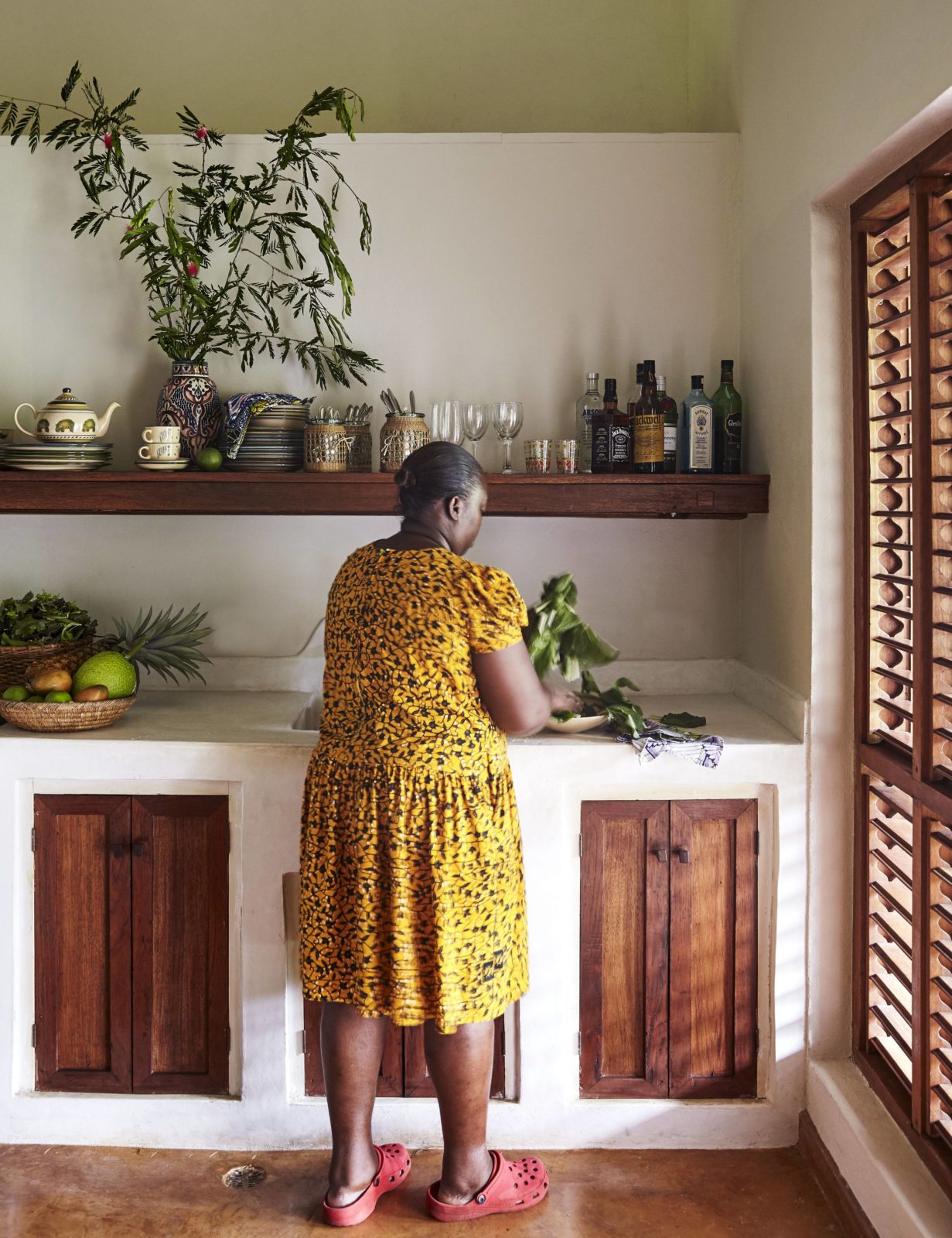 An indoor shot of a woman in a yellow leopard-print dress standing in a rustic kitchen, preparing green leafy vegetables on a white countertop. The kitchen features dark wooden cabinet doors and a floating wooden shelf with bottles and dishware.