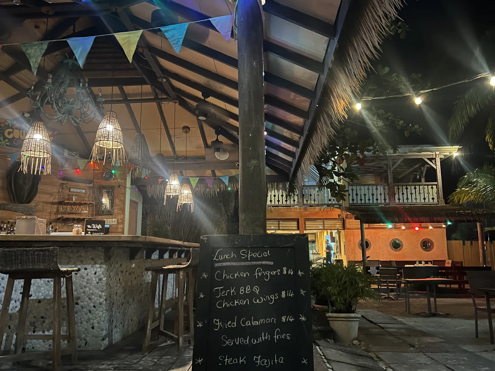open-air bar at night with woven pendants, chalkboard specials and festoon lights, verandah beyond.