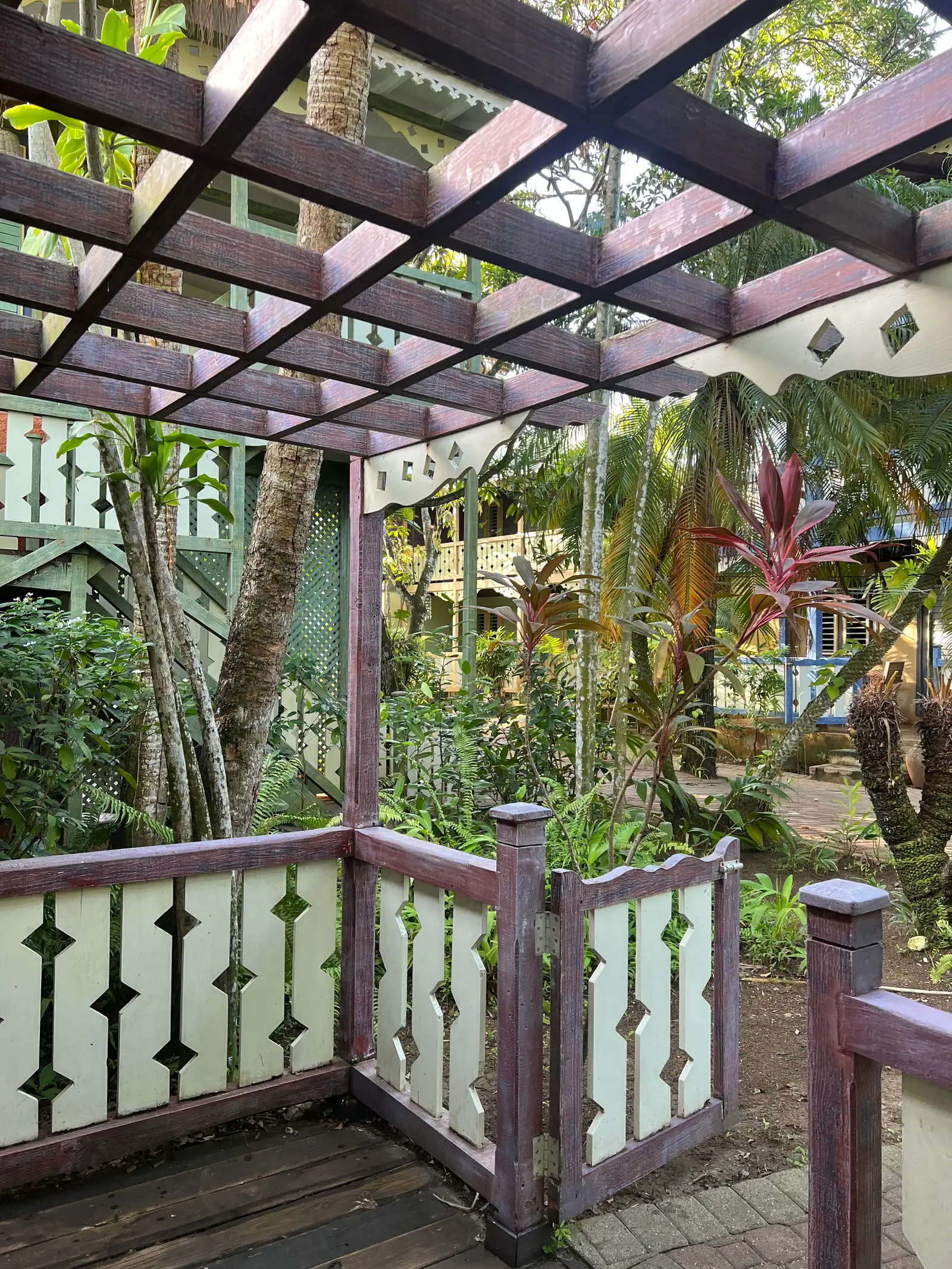 painted timber pergola and small gate with diamond cutouts, cottages and palms beyond.