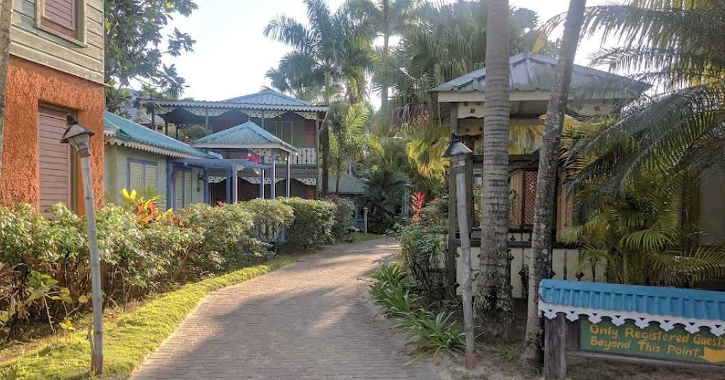 A brick-paved driveway leading through a tropical garden complex of traditional Jamaican cottages. The colorful wooden cottages feature teal and orange accents, intricate wooden railings, and are surrounded by high hedges and dense palm trees. A small sign for "Only Registered Guests" is visible in the foreground.