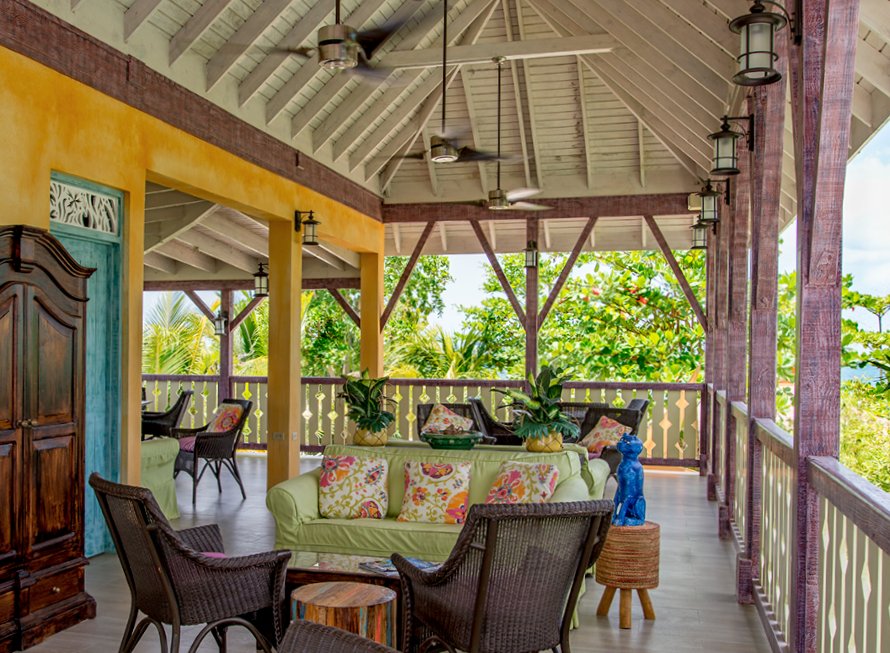 A colorful, covered porch or veranda featuring traditional Caribbean architecture with white slatted ceilings and pink/purple timber framing. The seating area includes a light green slipcovered sofa with colorful floral pillows, woven wicker chairs, and a dark carved wooden cabinet against a yellow stucco wall.