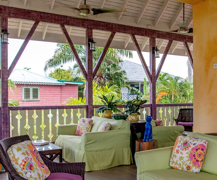 A vibrant outdoor porch area with light green slipcovered sofas and wicker chairs, featuring pillows with colorful floral patterns. The porch has a dark purple/red timber frame with diagonal beams and hanging lanterns, overlooking a lush tropical garden with a pink wooden cottage in the background.