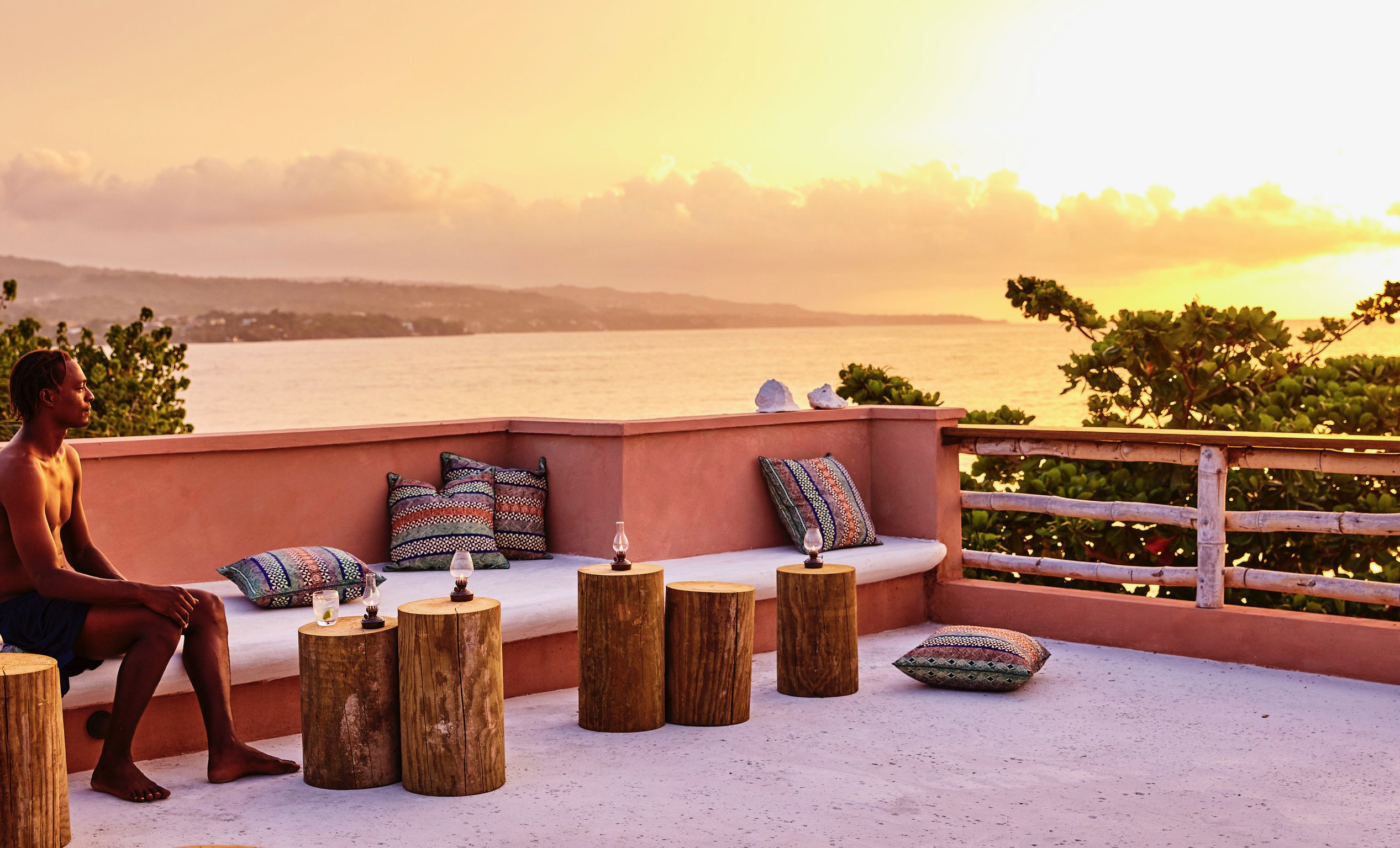 rooftop daybed beneath a bamboo pergola, guest reclining with a bottle, terracotta parapet and village roofs against low hills.