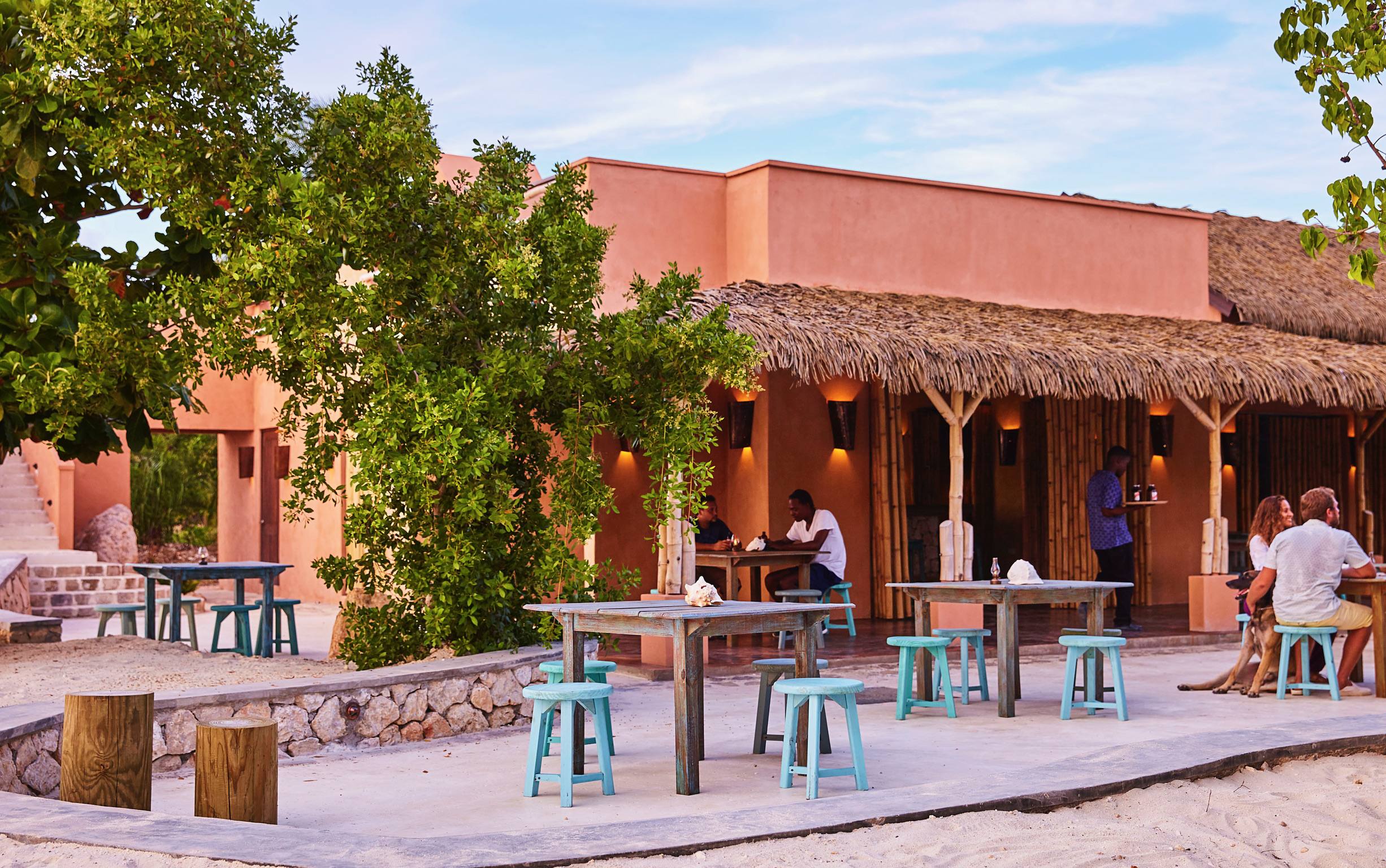 An outdoor dining area on a sandy patio in front of a building with salmon-pink walls and a thatched roof. Light blue wooden tables and stools are scattered, with people dining in the background.