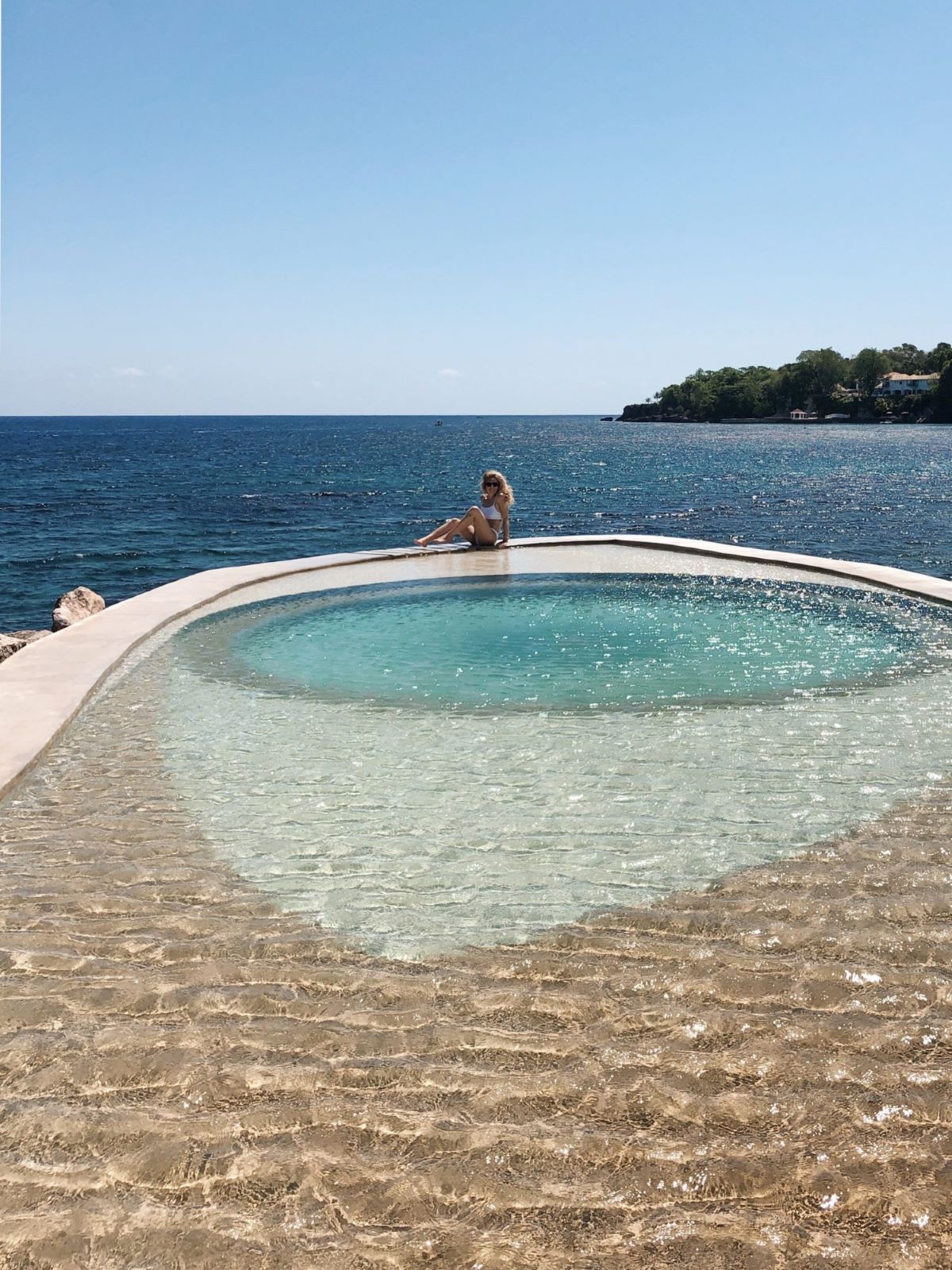 a curved, tiered infinity pool, looking out over the clear blue ocean under a bright, clear sky. The pool's edge meets the sea, and the water in the pool transitions from a deep blue at the back to a light, shimmering golden color in the foreground due to the sun and shallow depth. A rocky coastline covered in greenery is visible in the distance to the right.
