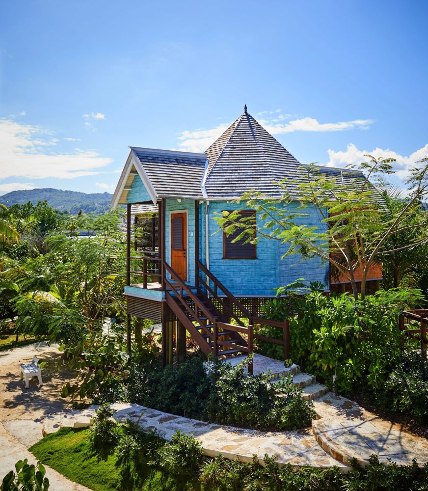 An elevated, light-blue wooden beach hut with a shingled roof is nestled among lush green tropical plants under a sunny blue sky. The hut is accessed by a set of dark wood stairs, and a winding stone path leads up to it from the foreground. Hills are visible in the background.