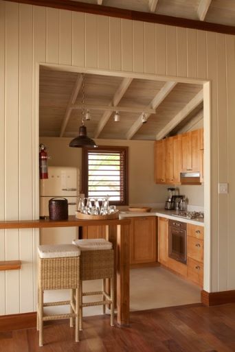 The interior view of a tropical-style kitchen visible through a serving bar opening. The kitchen has light wood cabinets, white appliances (including a vintage-style refrigerator), and a vaulted ceiling with exposed white beams. Wicker-seated bar stools are placed at the counter in the foreground.