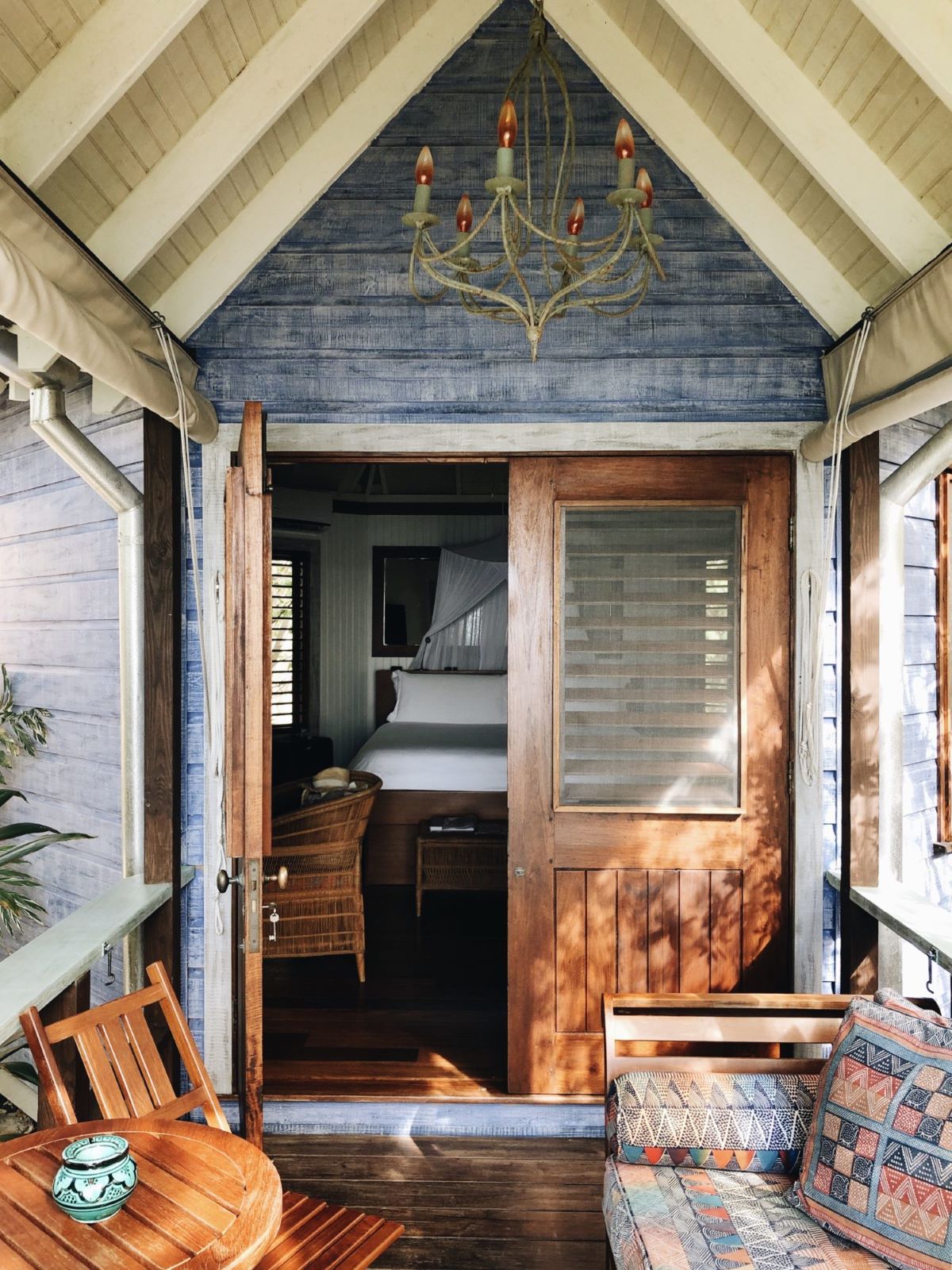 A warm, inviting close-up view of a cottage's covered porch entrance. The exterior walls are painted a weathered blue, and the A-frame ceiling is white and blue. Wooden double doors are open, revealing a bedroom with a canopied white bed inside. On the porch, there's a wooden table with a small teal bowl, a wicker chair, and a built-in bench seat with colorful, patterned cushions. A rustic metal chandelier hangs overhead.
