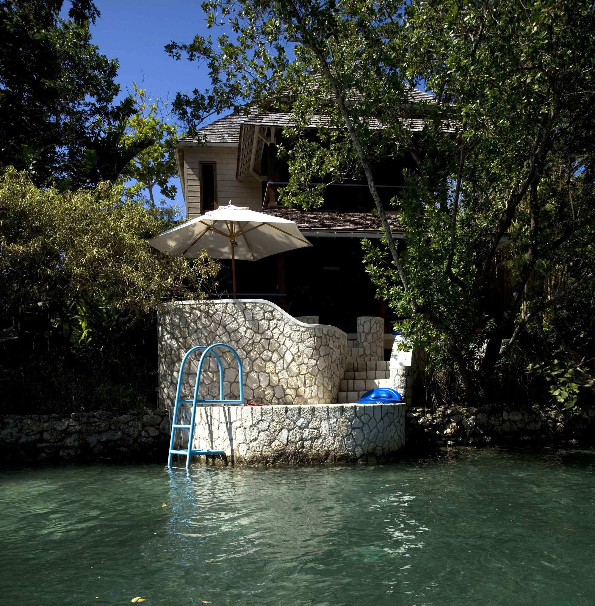 A two-story wooden cottage, nestled behind dense green trees, overlooks a clear, turquoise-green body of water. A white stone wall with curved steps and a small landing extends from the cottage into the water. A bright blue ladder is anchored to the stone wall, providing access for swimming. A white sun umbrella is open on the patio above the steps.