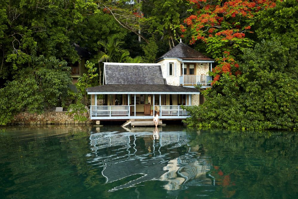 A tranquil wooden cottage spa building, painted light blue and white with a dark roof, is situated at the edge of a calm, dark turquoise body of water, possibly a lagoon or bay. The building is surrounded by dense, vibrant green foliage and trees with patches of red-orange flowers. A woman sits on the small wooden dock, and the cottage's reflection shimmers on the water's surface.
