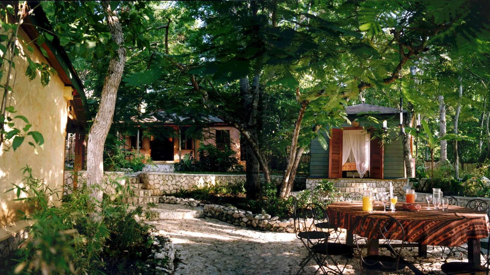 shaded garden courtyard with stone paths and outdoor dining table, timber cottages around and an open shutter revealing a mosquito-net bed.