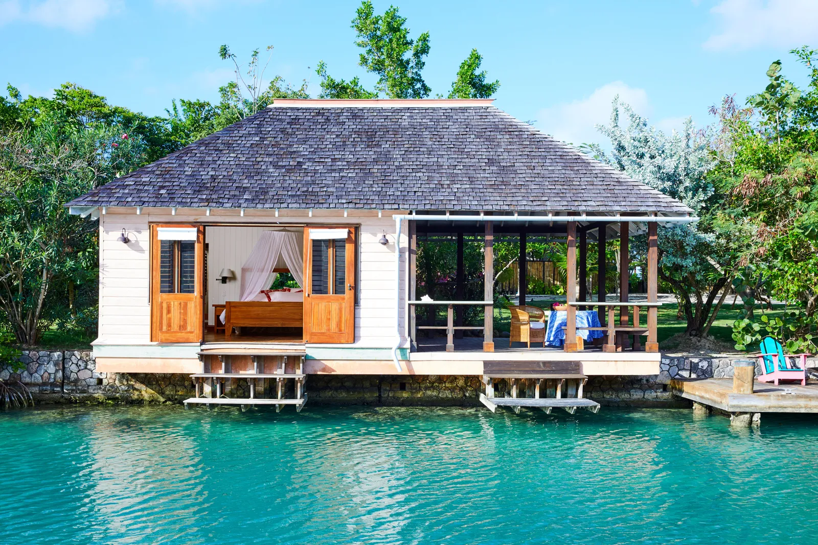 lagoon cottage on stilts over turquoise water, doors open to a four-poster bed with netting and a verandah seating area.