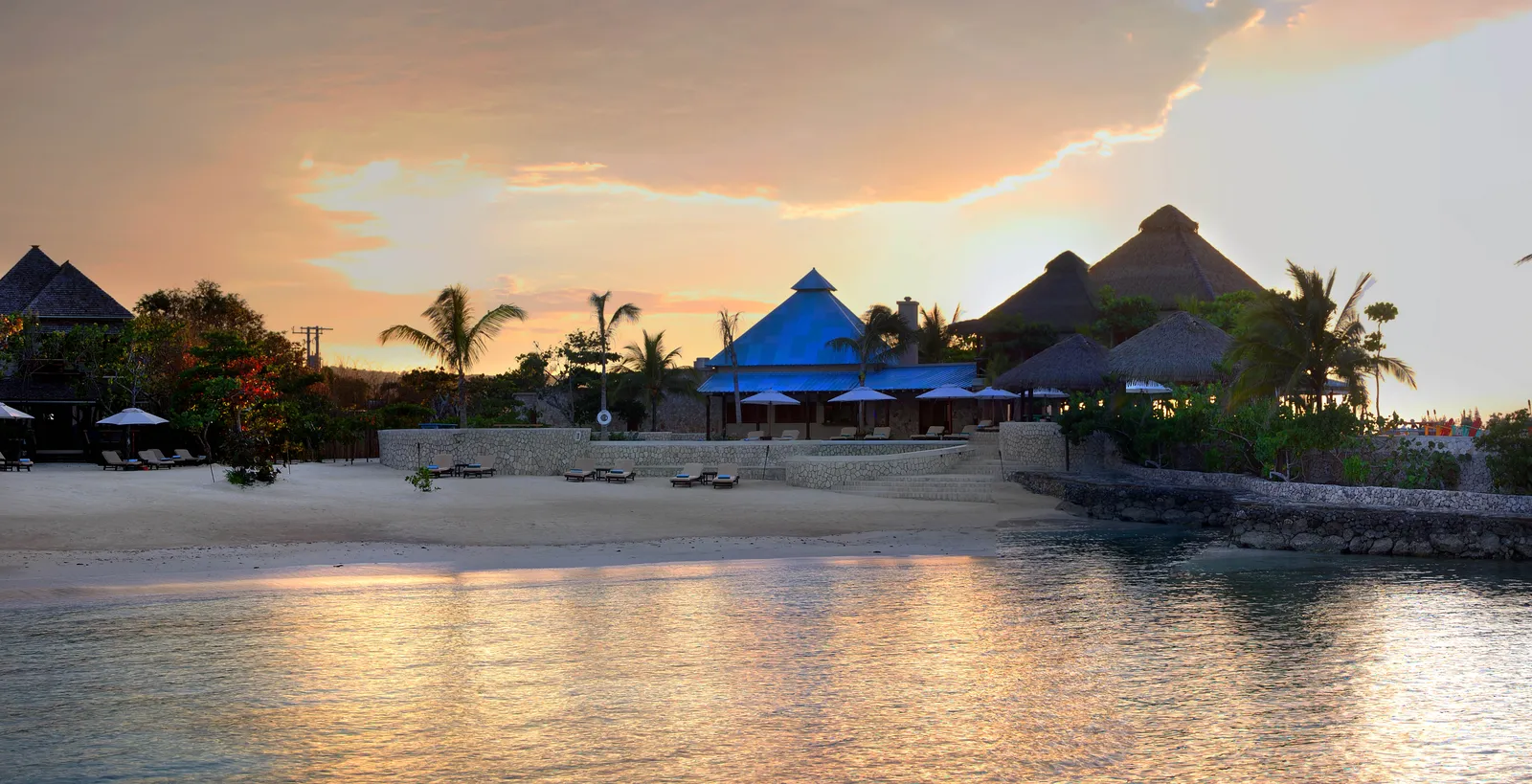 sunset over the resort’s sandy cove, blue-roofed pavilion and thatch huts with palms and umbrellas reflected in calm water.