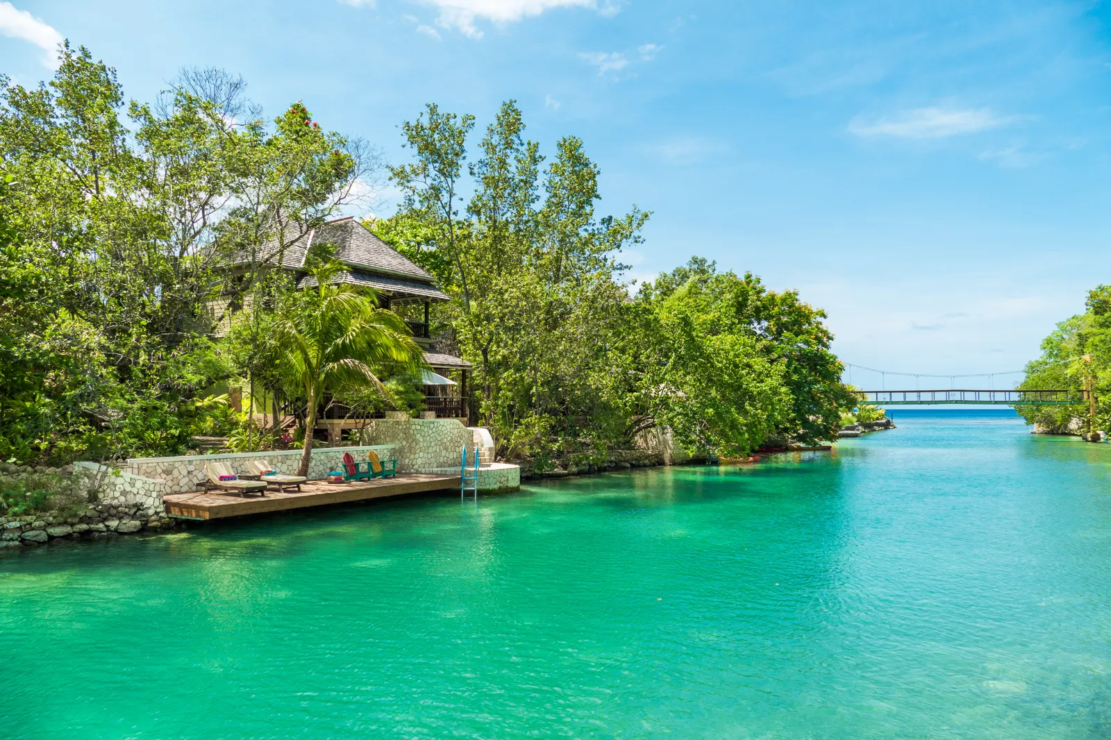 quiet lagoon channel with stone quay, colorful deck chairs and trees, opening to the sea with the bridge in the distance.