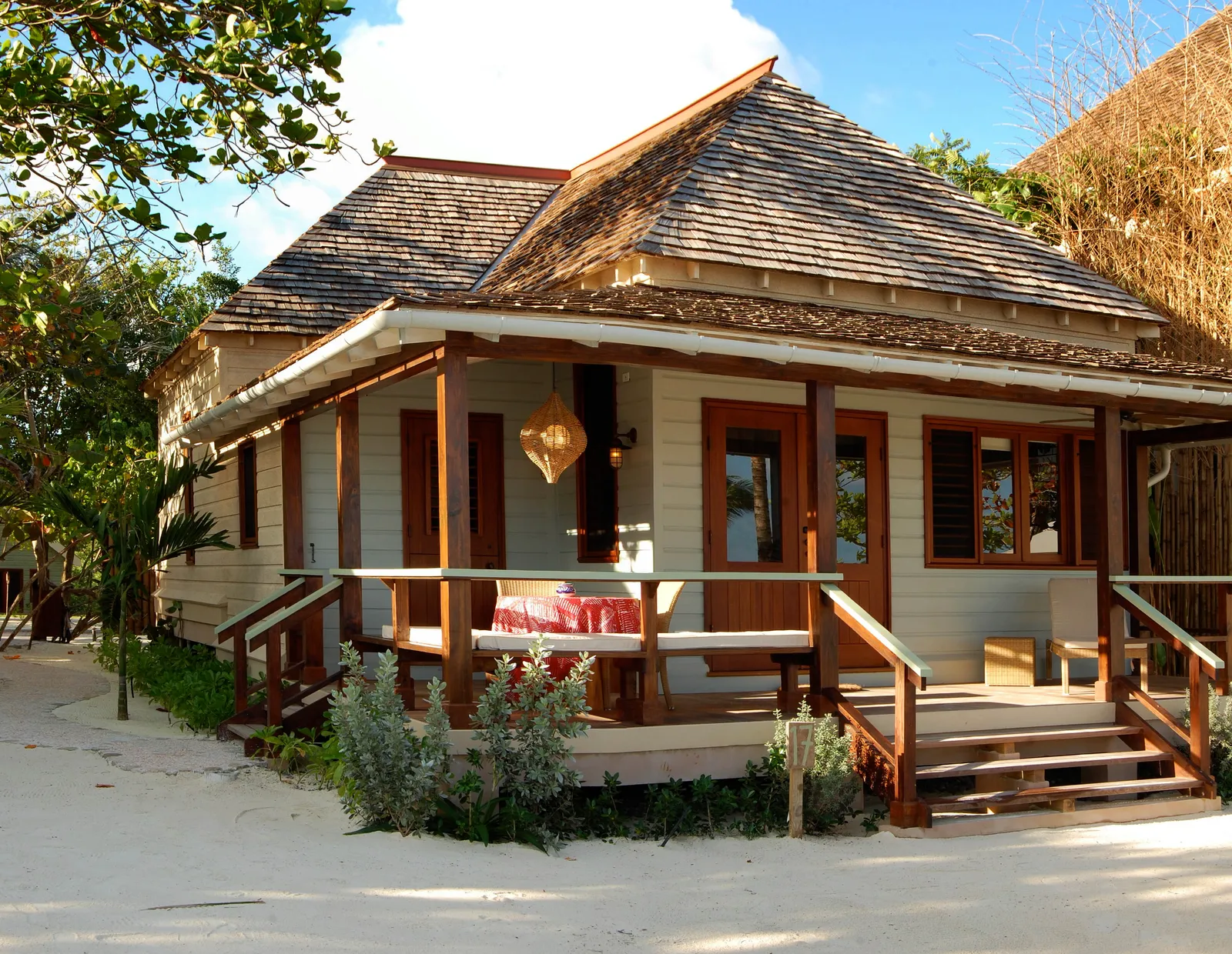 single-storey beach cottage with wraparound verandah and timber posts, steps down to white sand and greenery.