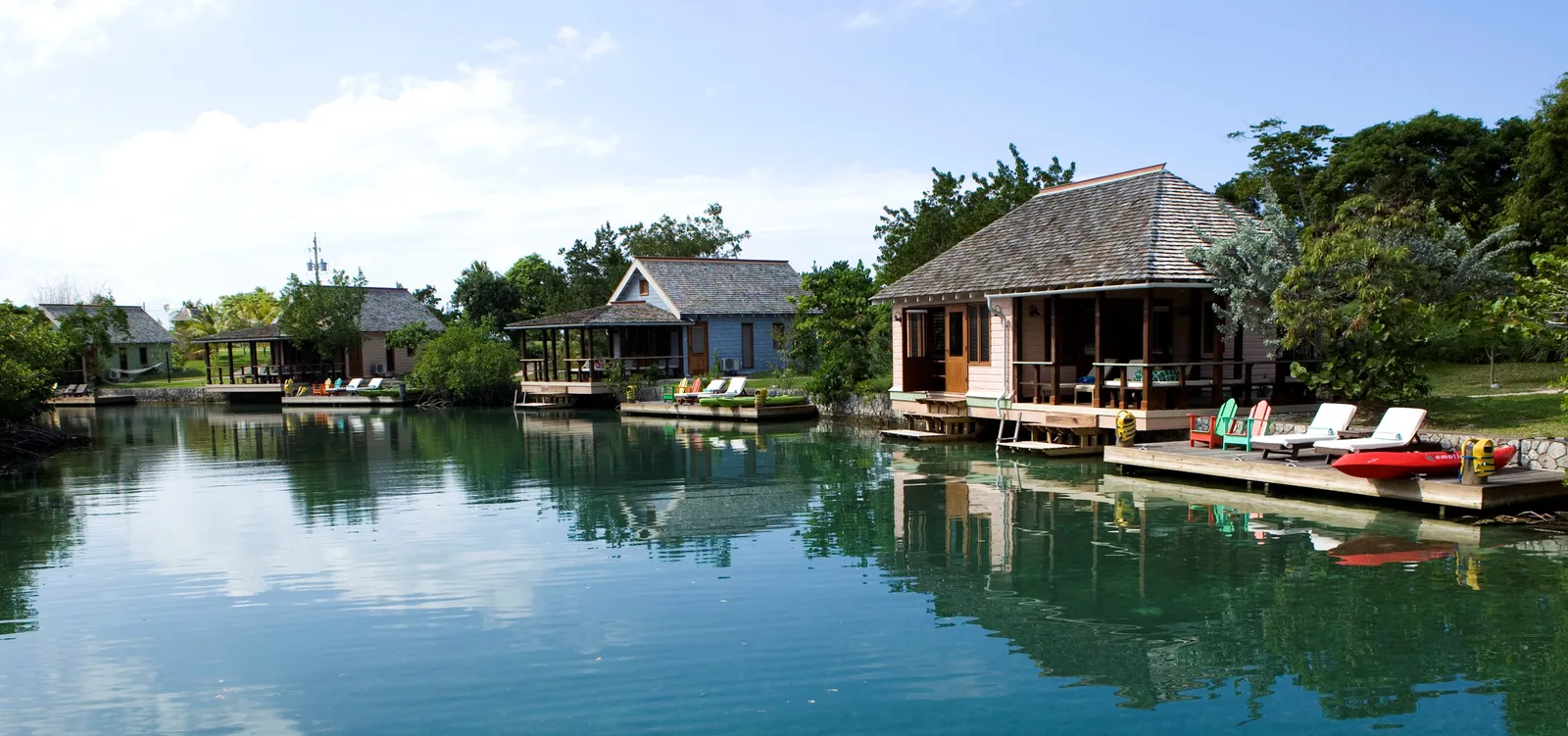 lagoon cottages on pilings with decks and sun loungers reflected in calm water, kayaks tied to a small dock.