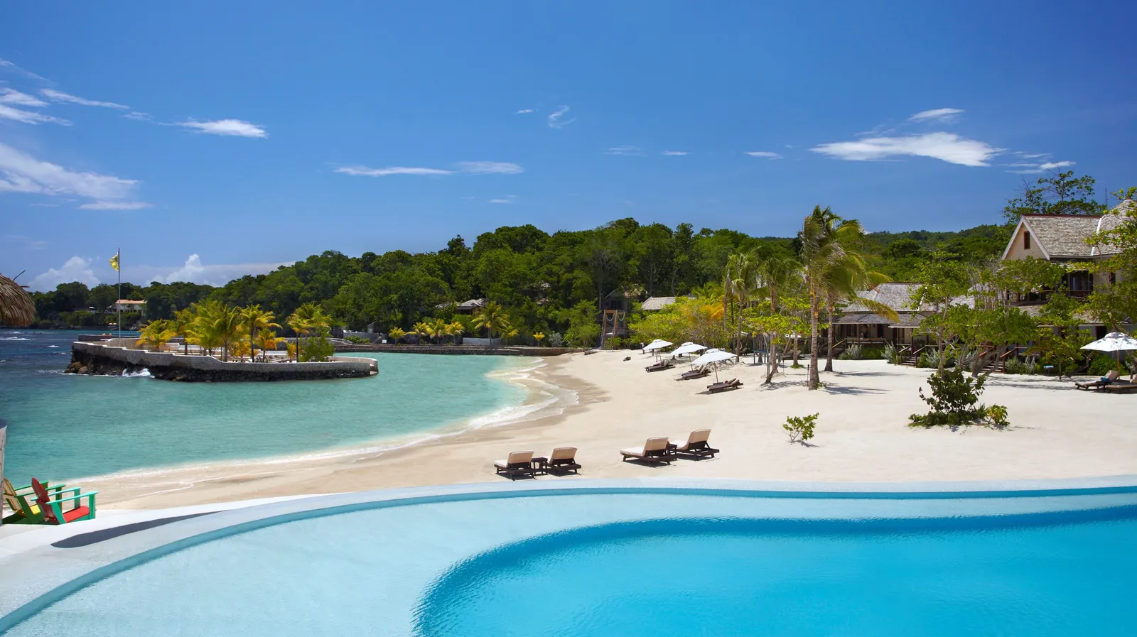 wide crescent beach and palm-lined breakwater seen beyond a curved infinity pool under a clear blue sky.