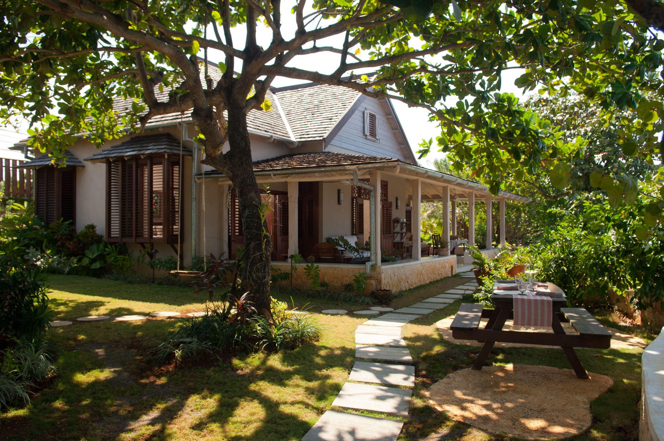 A lush tropical garden area with mature trees framing the view of the ocean, a curved stone path with stepping stones leading past the corner of the house and a veranda, with a small pool or jacuzzi visible near the cliff edge.