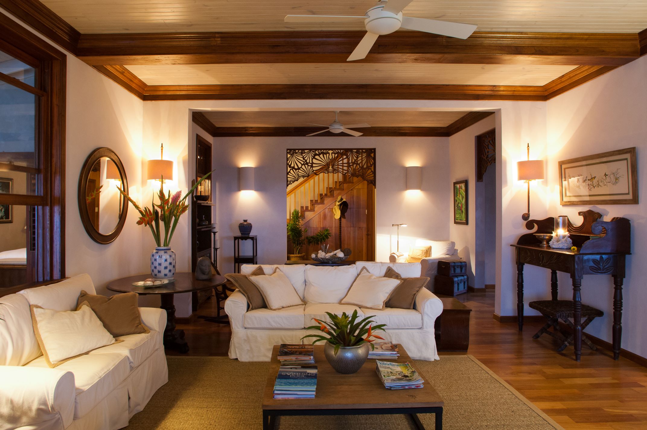 A warmly lit, inviting living room with two white slipcovered sofas, a central wood coffee table, and hardwood floors. The room features dark wood ceiling beams, wall sconces, and a view into an entryway with a winding staircase railing visible.