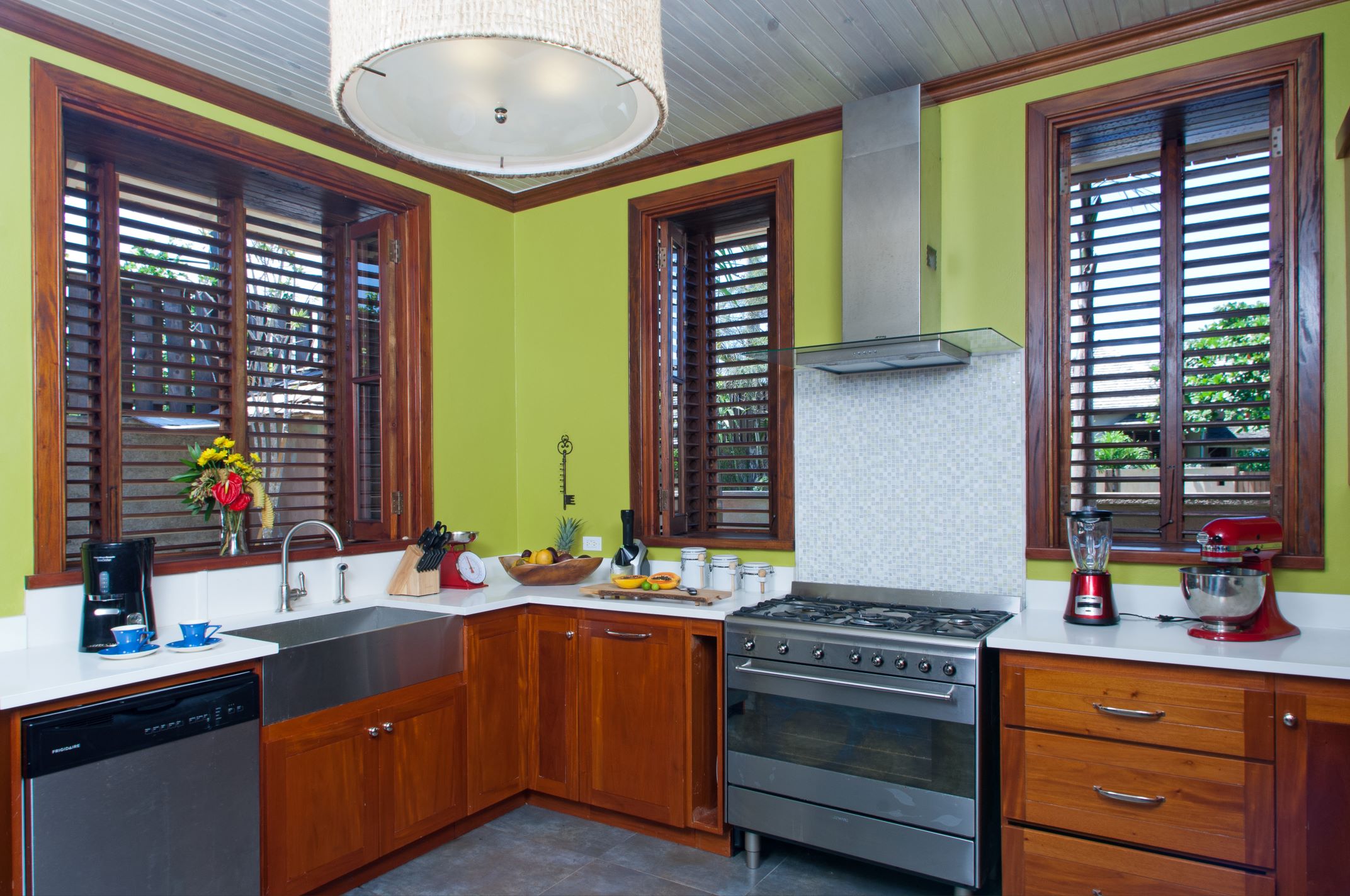 A bright kitchen with lime-green walls, white countertops, and warm-toned wood cabinetry and window frames, featuring a farmhouse sink, stainless steel appliances, and large windows with wooden shutters.