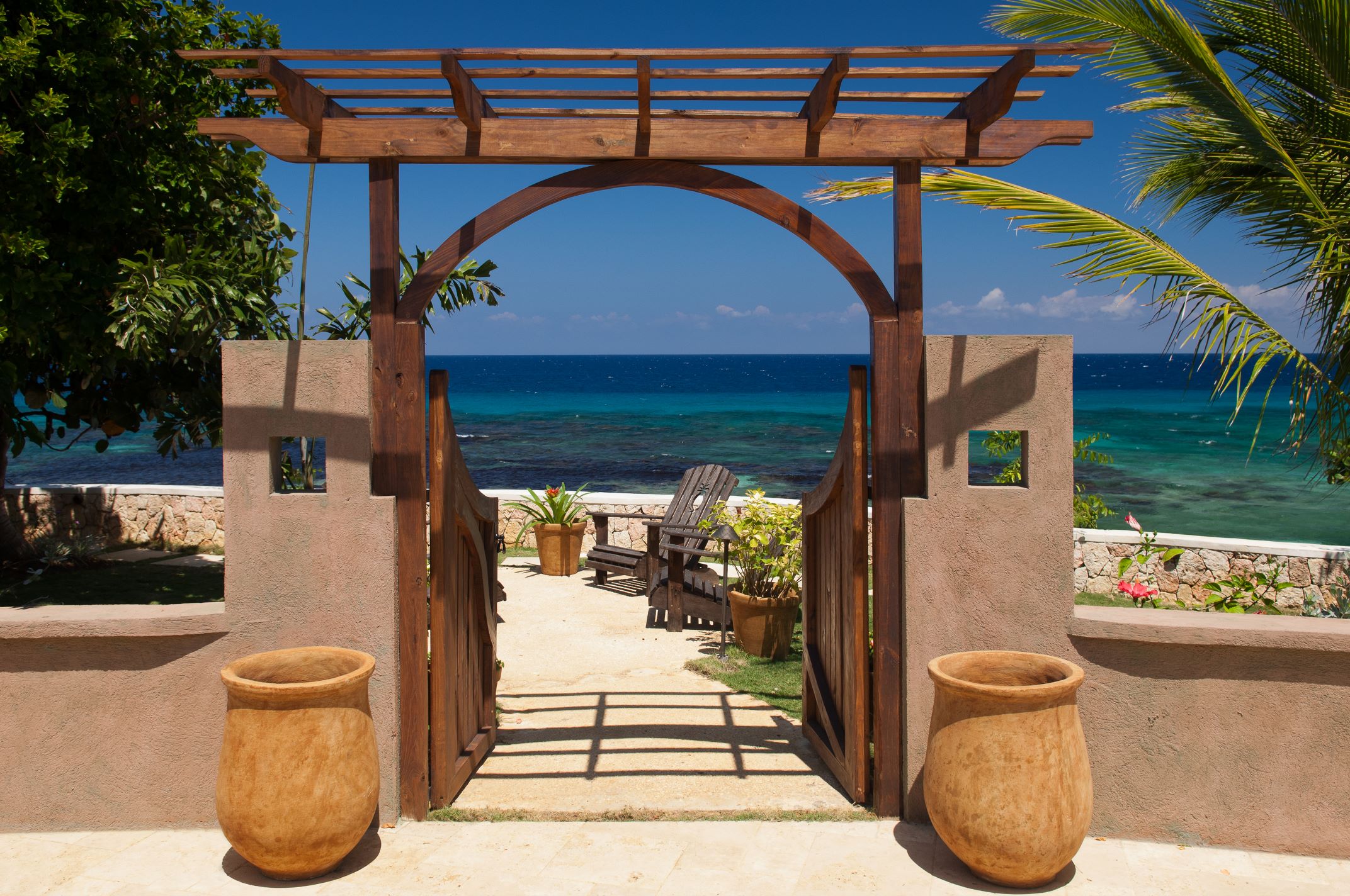 A rustic wooden arched gate with a lattice pergola at the top, set between two stucco walls, flanked by two large clay pots, opening to a view of the ocean, a deck, and an Adirondack chair.