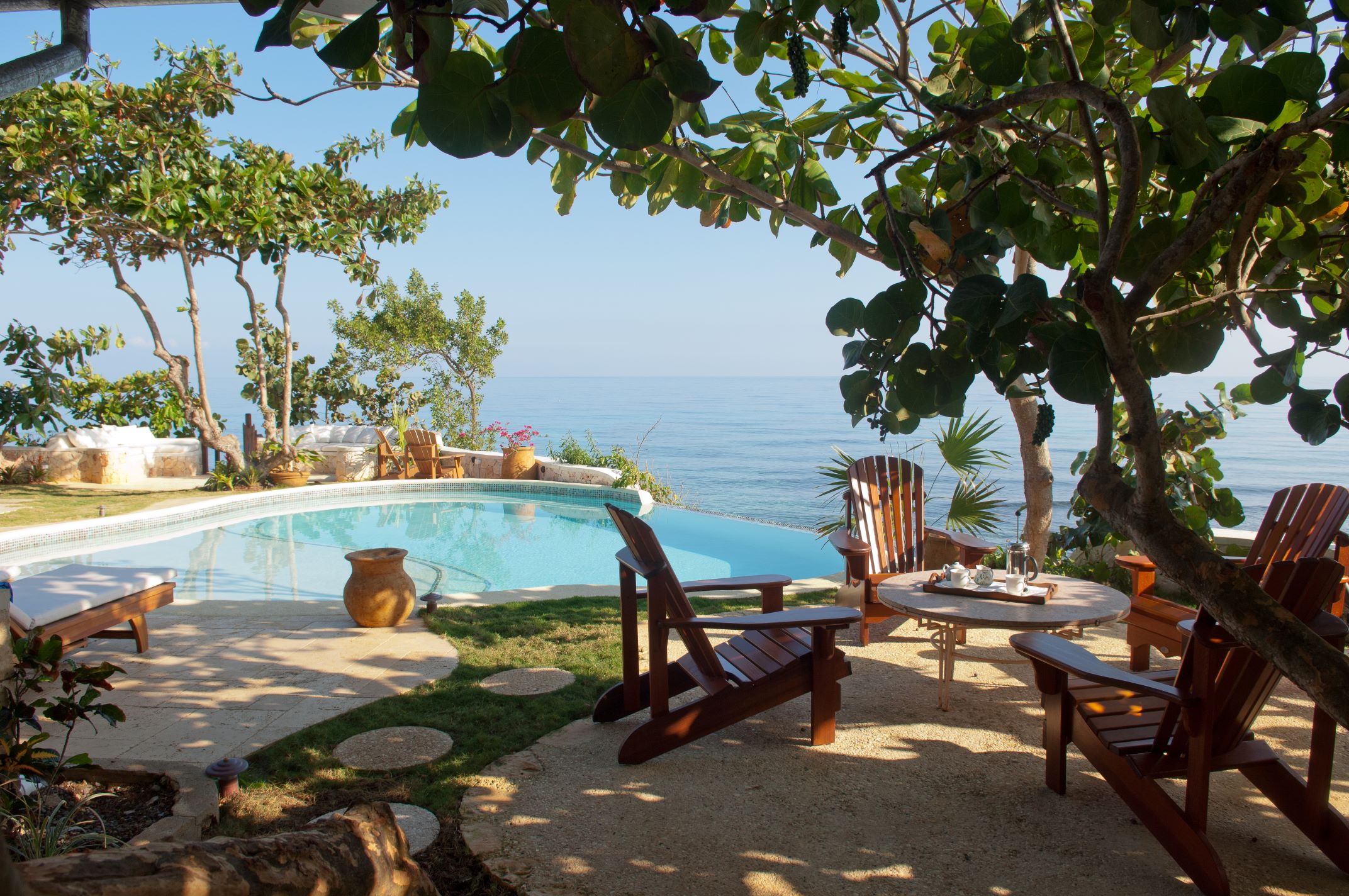 A shaded poolside area featuring dark wooden Adirondack chairs and a small round table with a drink setting, a terracotta vase near the water's edge, all framed by tropical trees and overlooking the clear blue ocean.