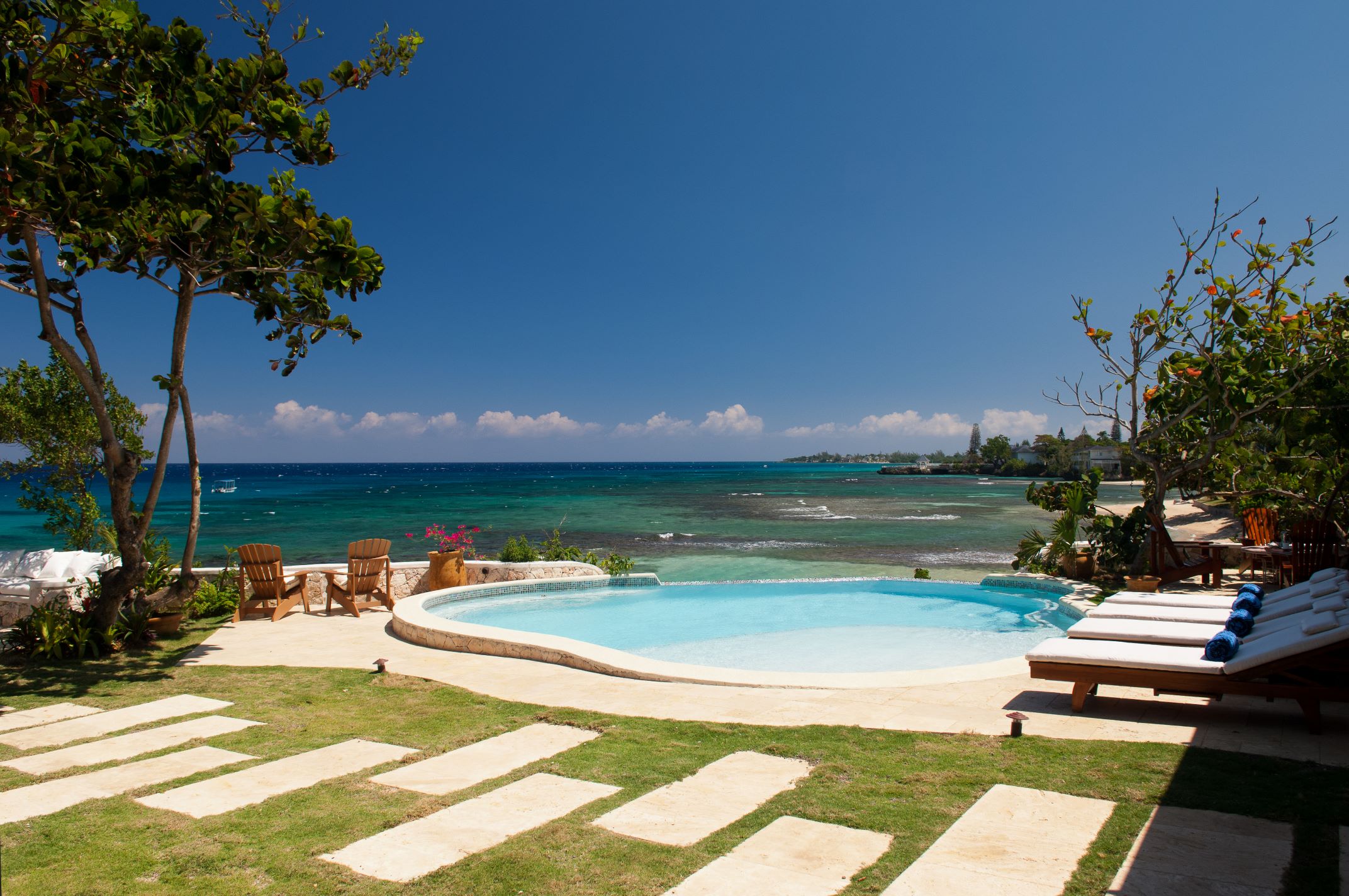 A curved, light blue swimming pool surrounded by a lawn with large stepping stones, wooden lounge chairs, and tropical trees, overlooking the turquoise and deep blue ocean under a clear sky.