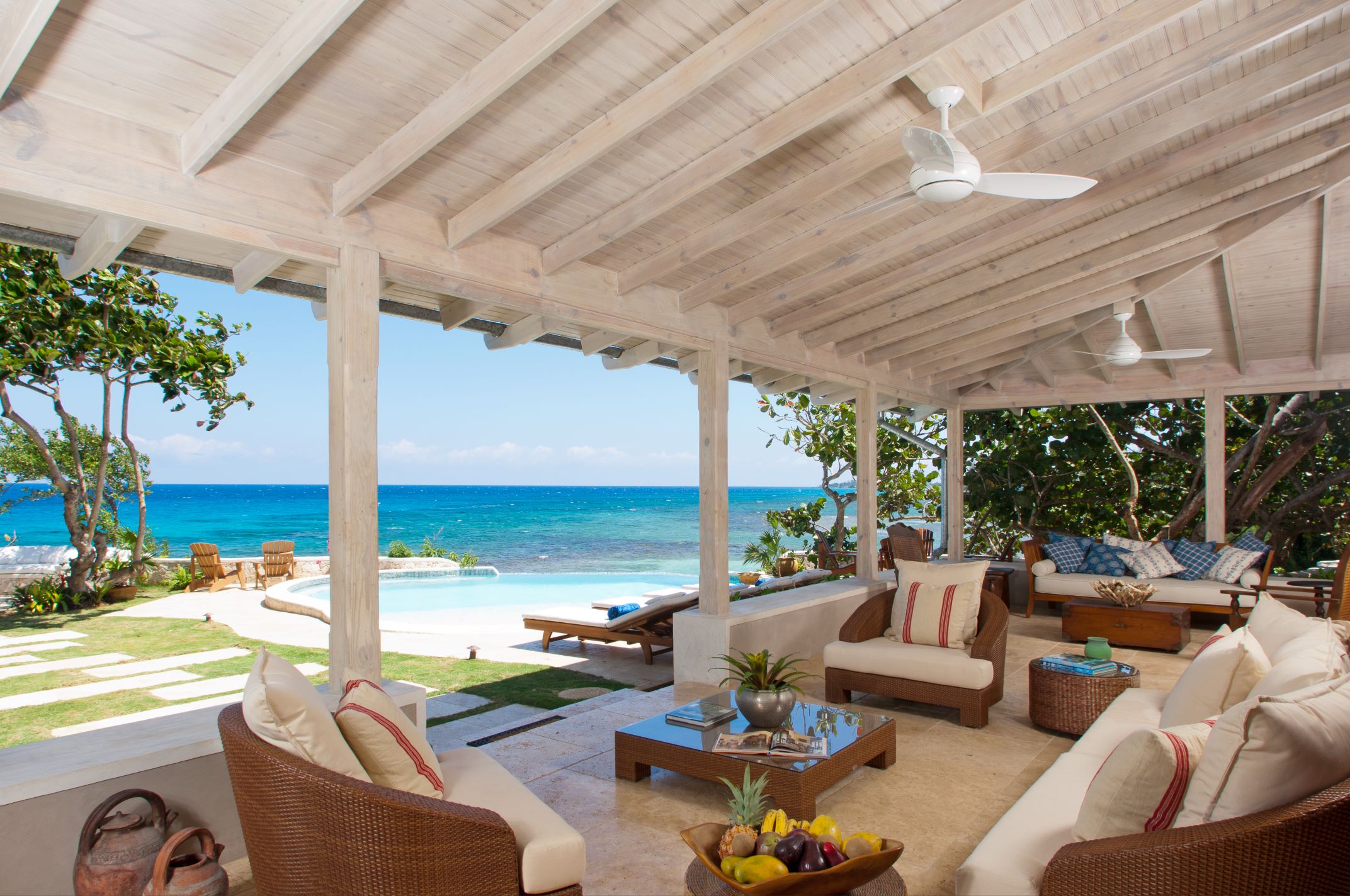 An open-air, covered patio with white-washed wood ceilings and columns, furnished with wicker and cushioned seating, a coffee table with fruit, all looking out onto a pool, lush lawn, and the bright blue ocean.