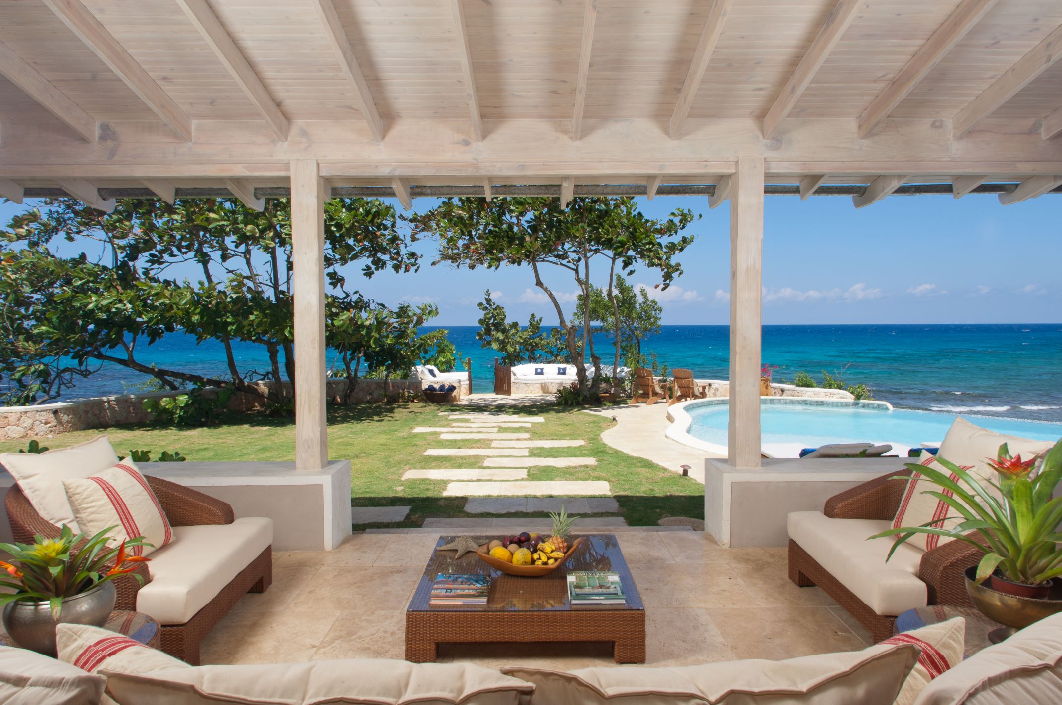 An outdoor covered veranda with comfortable wicker furniture and cushions, featuring a coffee table with a bowl of fruit. The view, framed by the white support posts, looks over a lawn with stepping stones, tropical trees, and a curved pool next to the ocean.