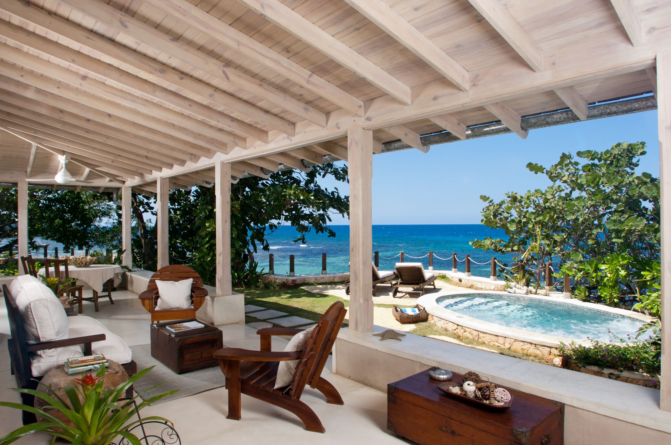 A covered patio with a white-washed plank ceiling and white-cushioned seating, including wooden Adirondack chairs, overlooking a lawn with a small, round hot tub or plunge pool and the deep blue ocean beyond a low fence.