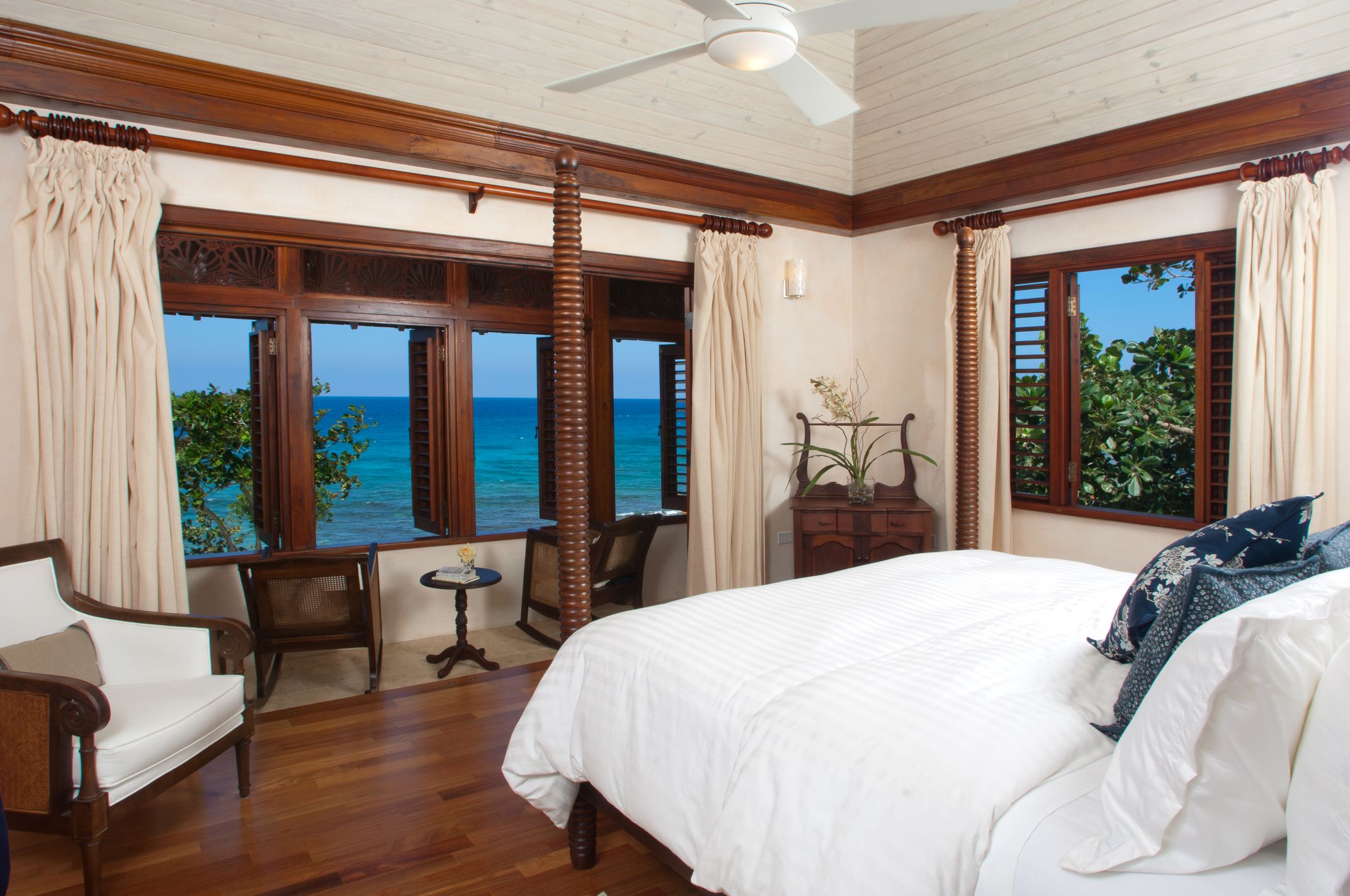 A bright bedroom with wood floors, a white bed, and wooden window frames featuring shutters. The windows open to an expansive view of the turquoise and deep blue ocean, with two rattan rocking chairs set in the middle window opening.