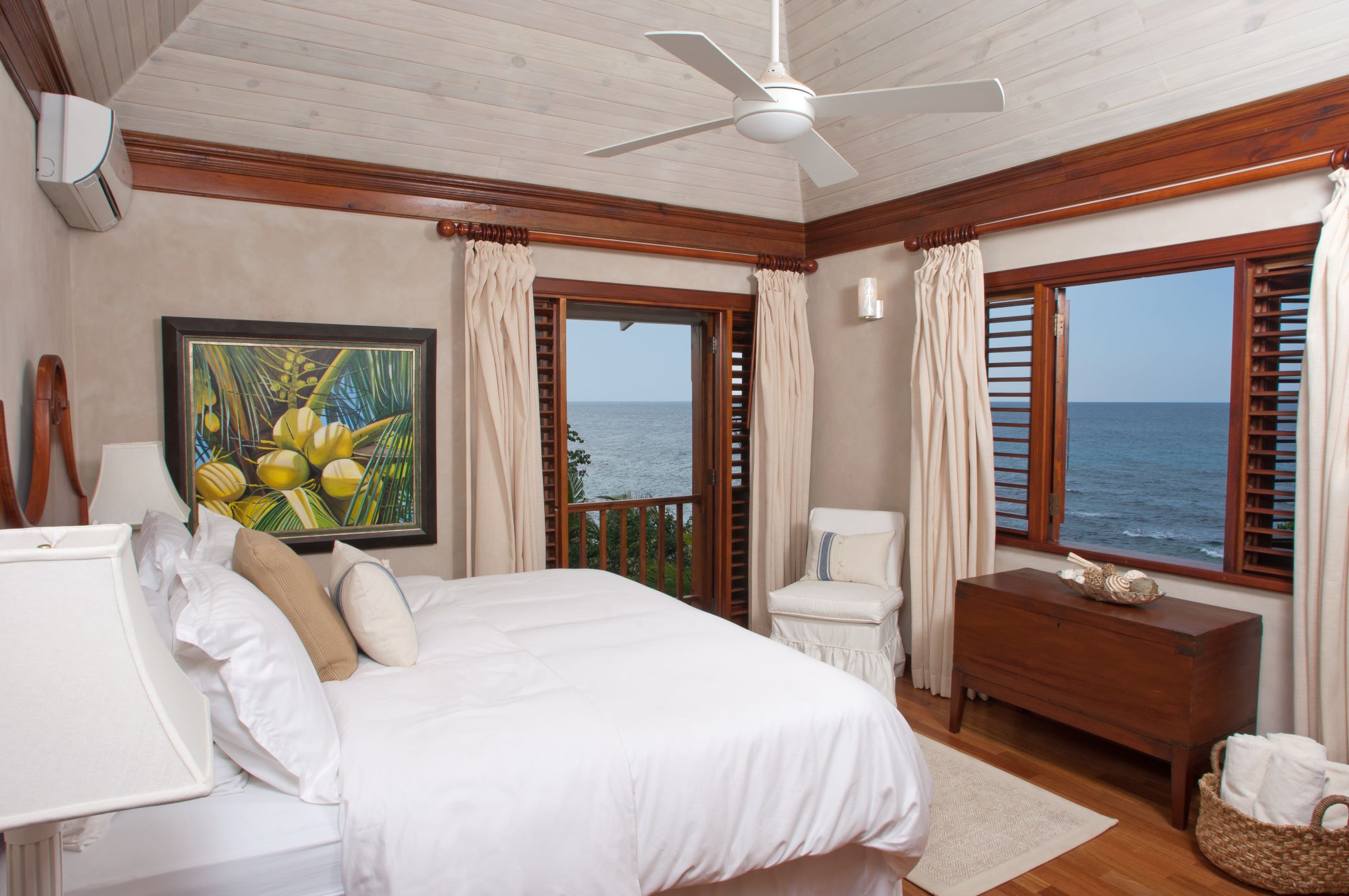 A bedroom featuring a white bed with light-colored pillows, rich dark wood trim, and a white-washed plank ceiling with a ceiling fan. The room has a window with wooden shutters and a door leading to a small balcony, both offering an ocean view, and a large painting of yellow coconuts on the wall.