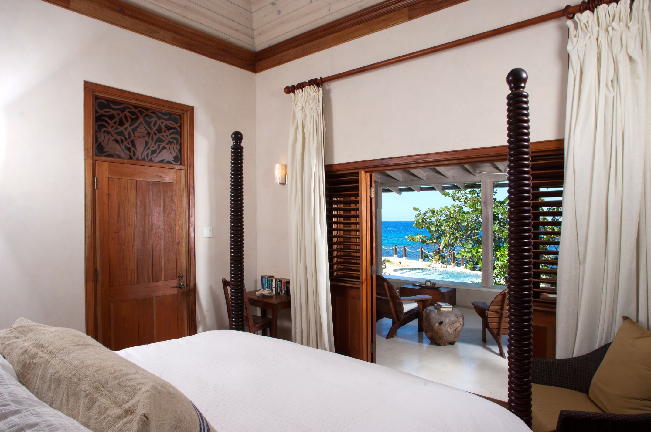 A bedroom with a four-poster bed and a white comforter, featuring dark wood trim and a unique carved wooden door on the left, and a large opening with wooden shutters that lead out to a private balcony with an ocean view.