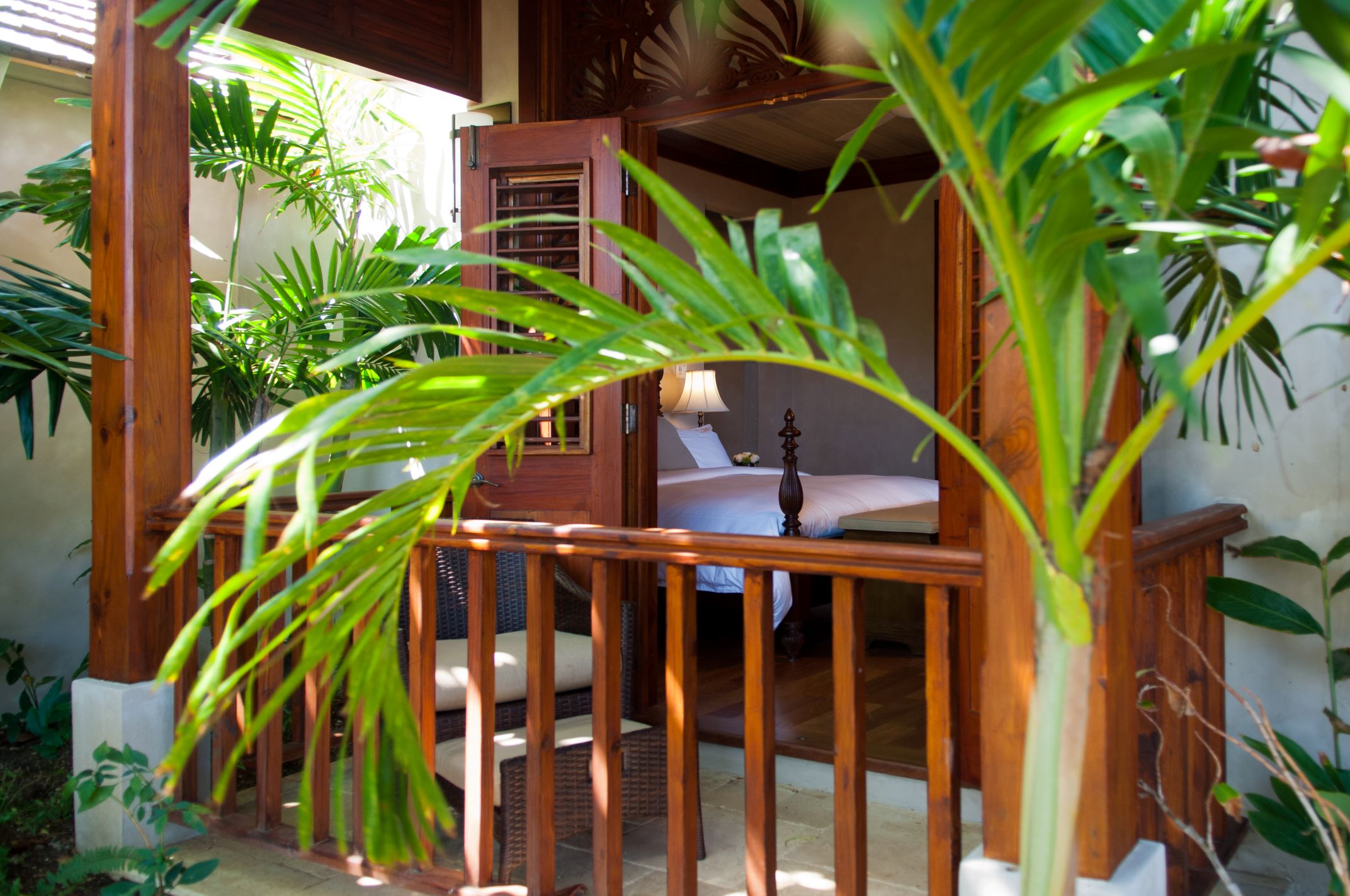 A close-up view through a wooden balcony door, framed by lush green palm fronds and wooden railings, revealing a glimpse of a sunlit bedroom with a white-covered bed inside.