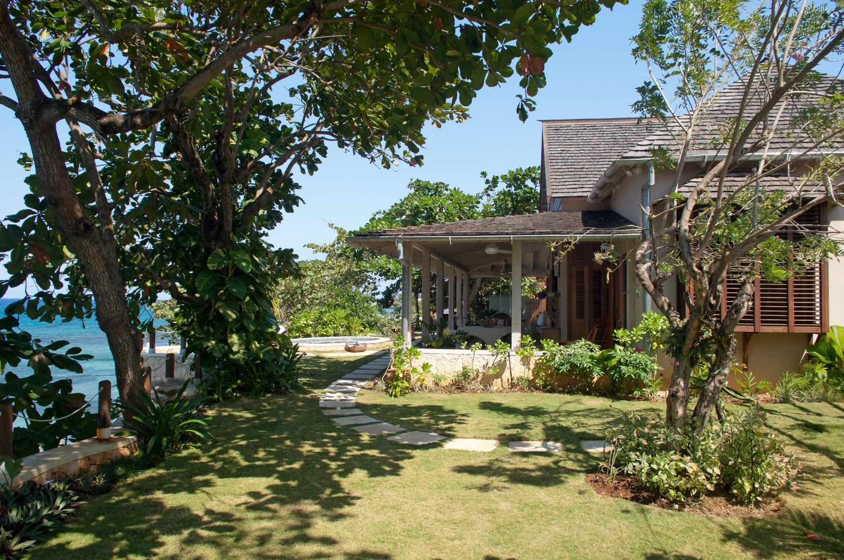 A lush tropical garden area with mature trees framing the view of the ocean, a curved stone path with stepping stones leading past the corner of the house and a veranda, with a small pool or jacuzzi visible near the cliff edge.