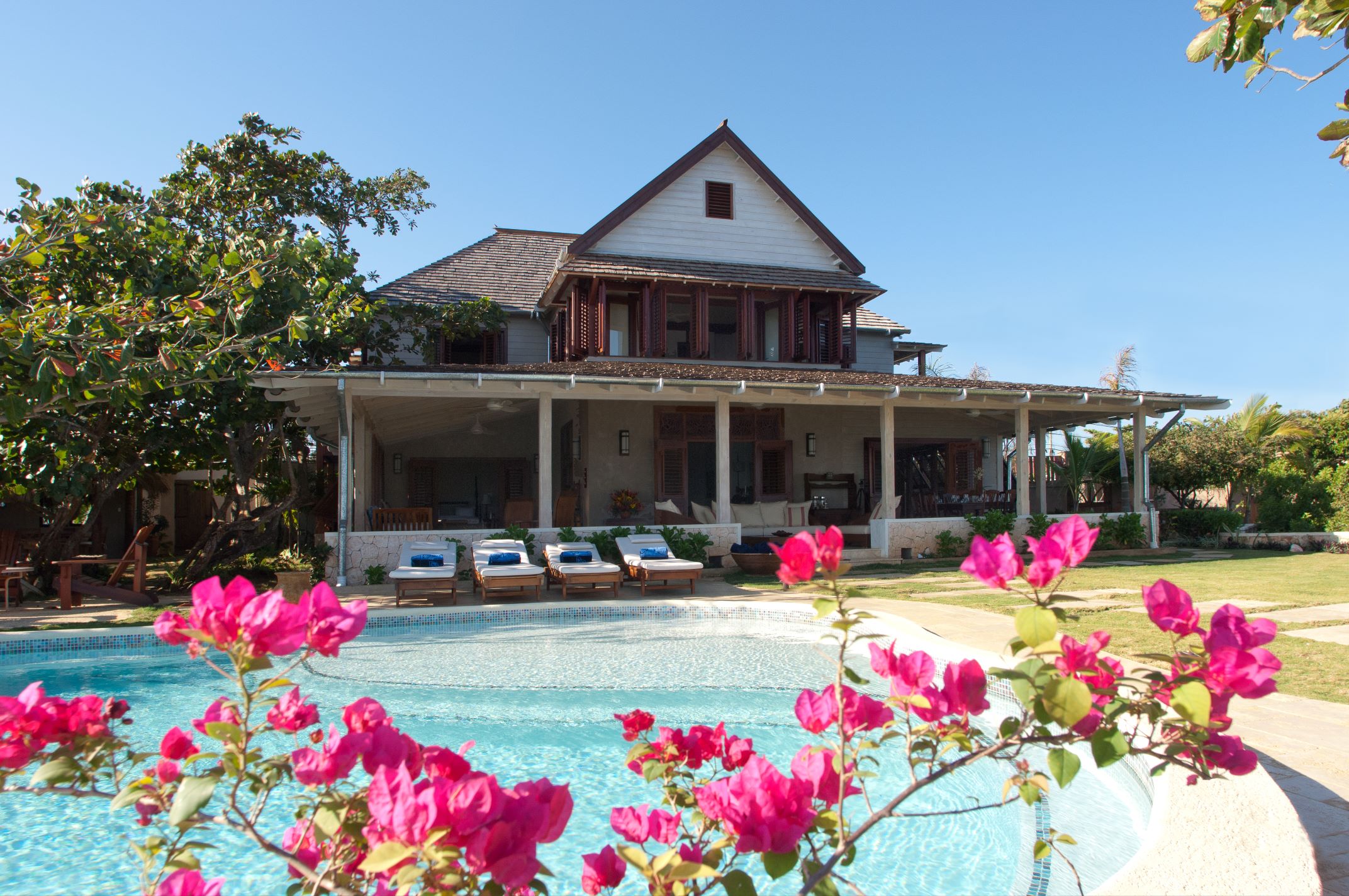 A grand, two-story vacation home or villa with a covered veranda and dark wood trim, set on a bright lawn behind a swimming pool, with vibrant pink bougainvillea flowers in the foreground.