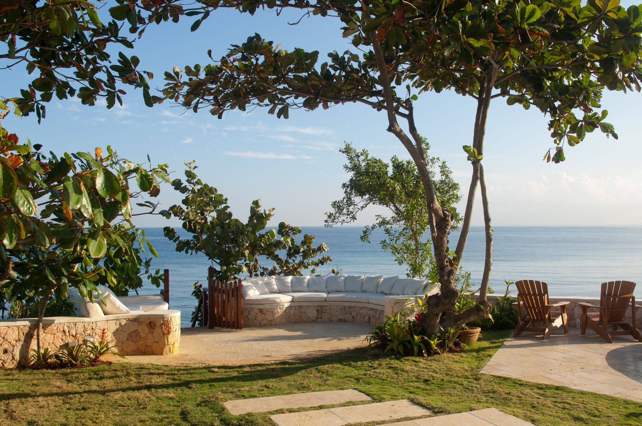 A sun-drenched outdoor patio area overlooking the ocean, featuring a built-in curved stone bench with white cushions and a set of wooden Adirondack chairs, all set on a stone deck under the shade of large tropical trees.