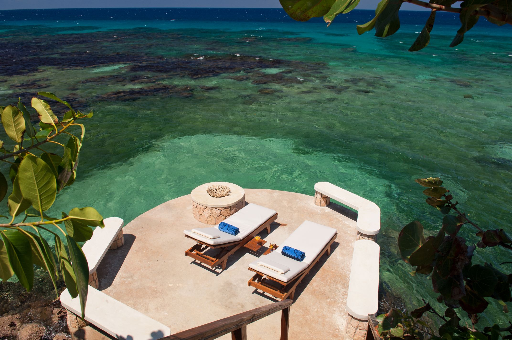 A circular, terracotta-colored deck built on a cliff edge with two wooden lounge chairs, a decorative stone fire pit, and built-in benches, providing a stunning view over the shallow, teal, and dark blue-green water.