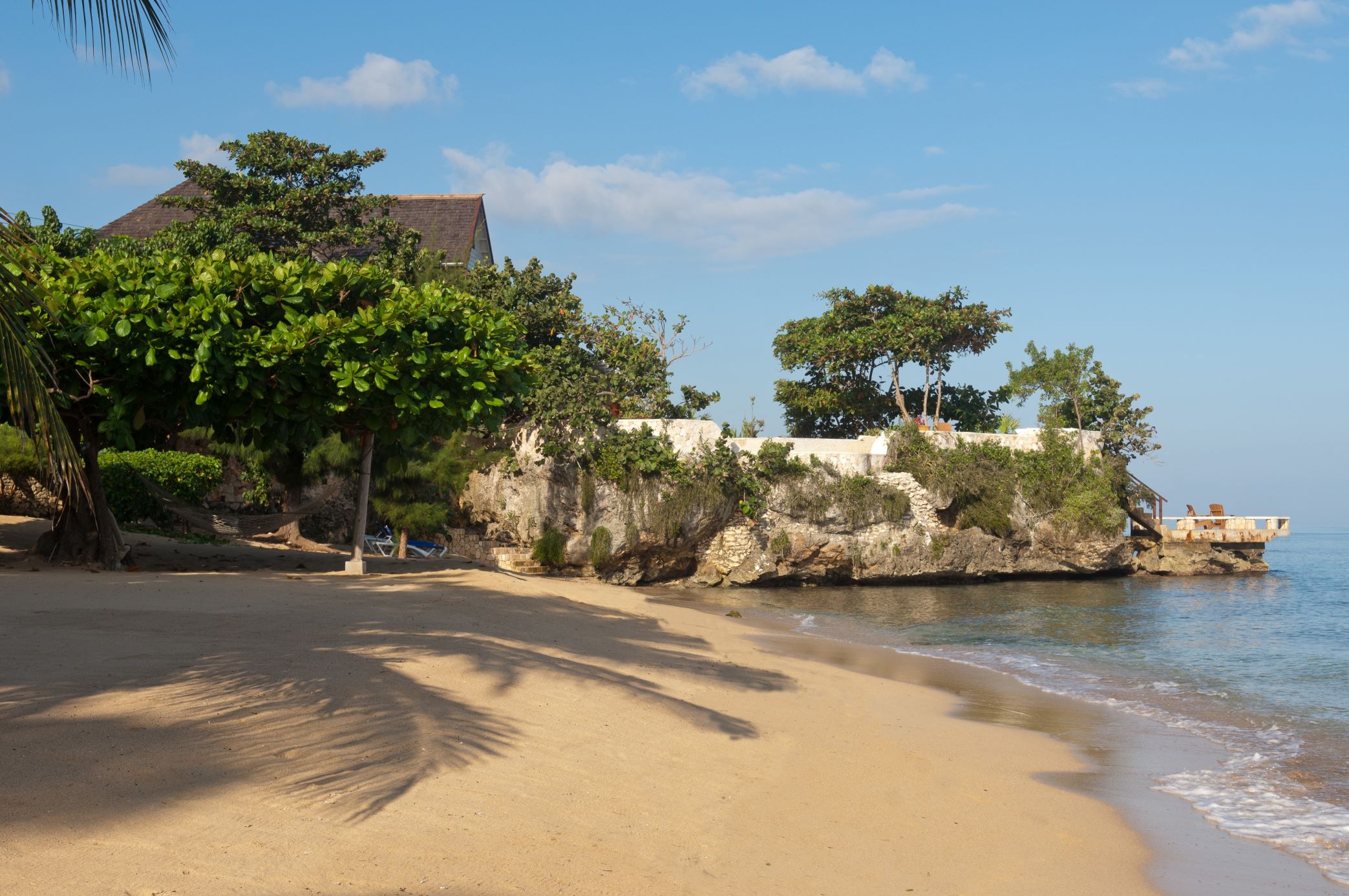 A wide shot of a secluded, sunlit sandy beach with gentle waves, palm tree shadows on the sand, and a rugged, vegetation-covered cliff or headland jutting into the water, topped with a structure.