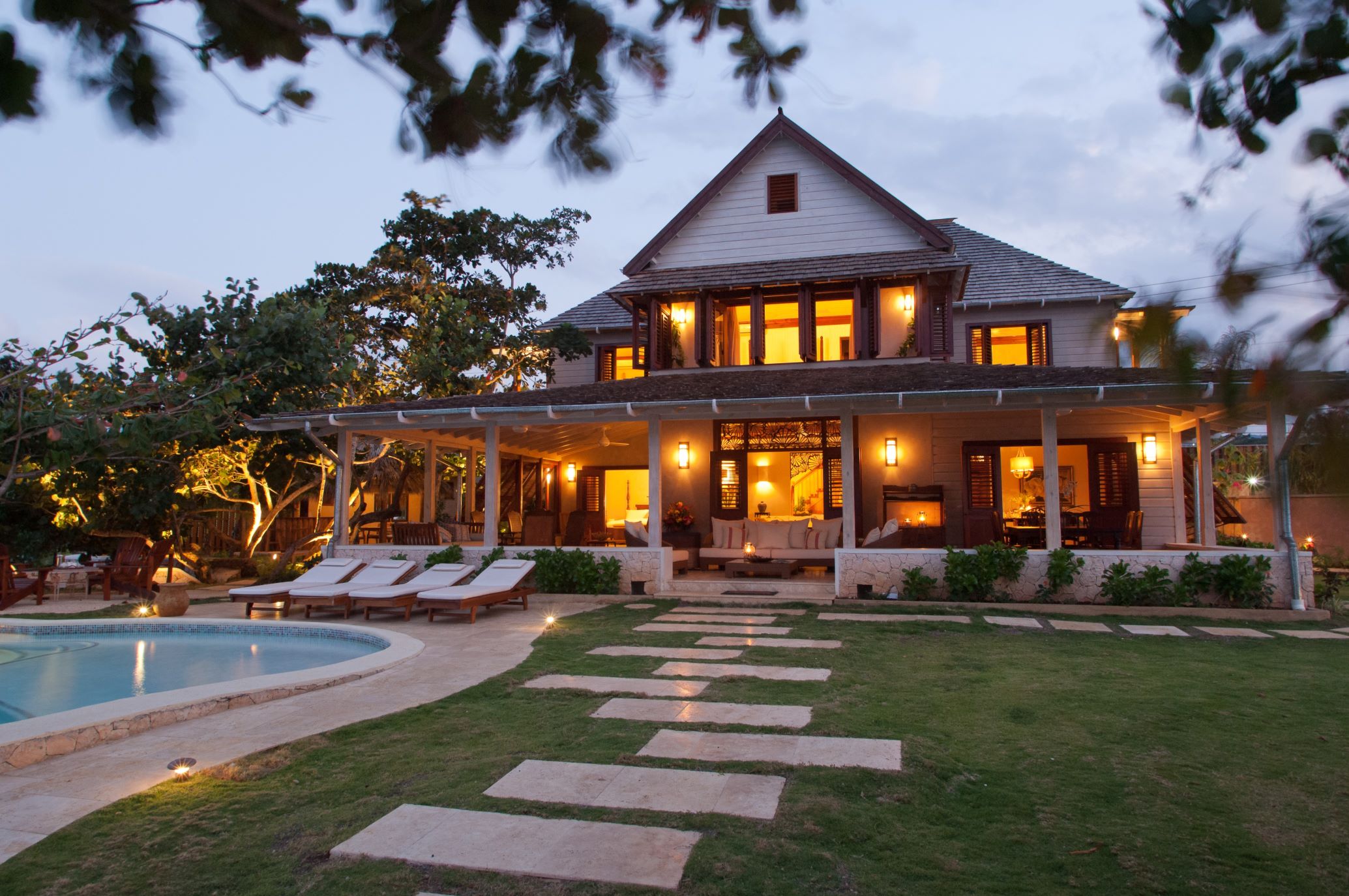 An evening exterior shot of a grand, two-story villa with a large pitched roof and a surrounding covered veranda, illuminated by warm interior and exterior lighting, next to a curved swimming pool with a stone path leading across the lawn.