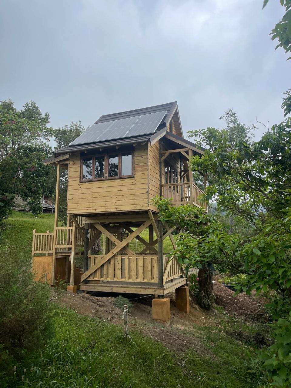 A newly constructed, elevated wooden tiny cabin at Holywell Park, Jamaica. The cabin features a small balcony, multi-pane windows, and is topped with solar panels, set on a grassy hill surrounded by trees.