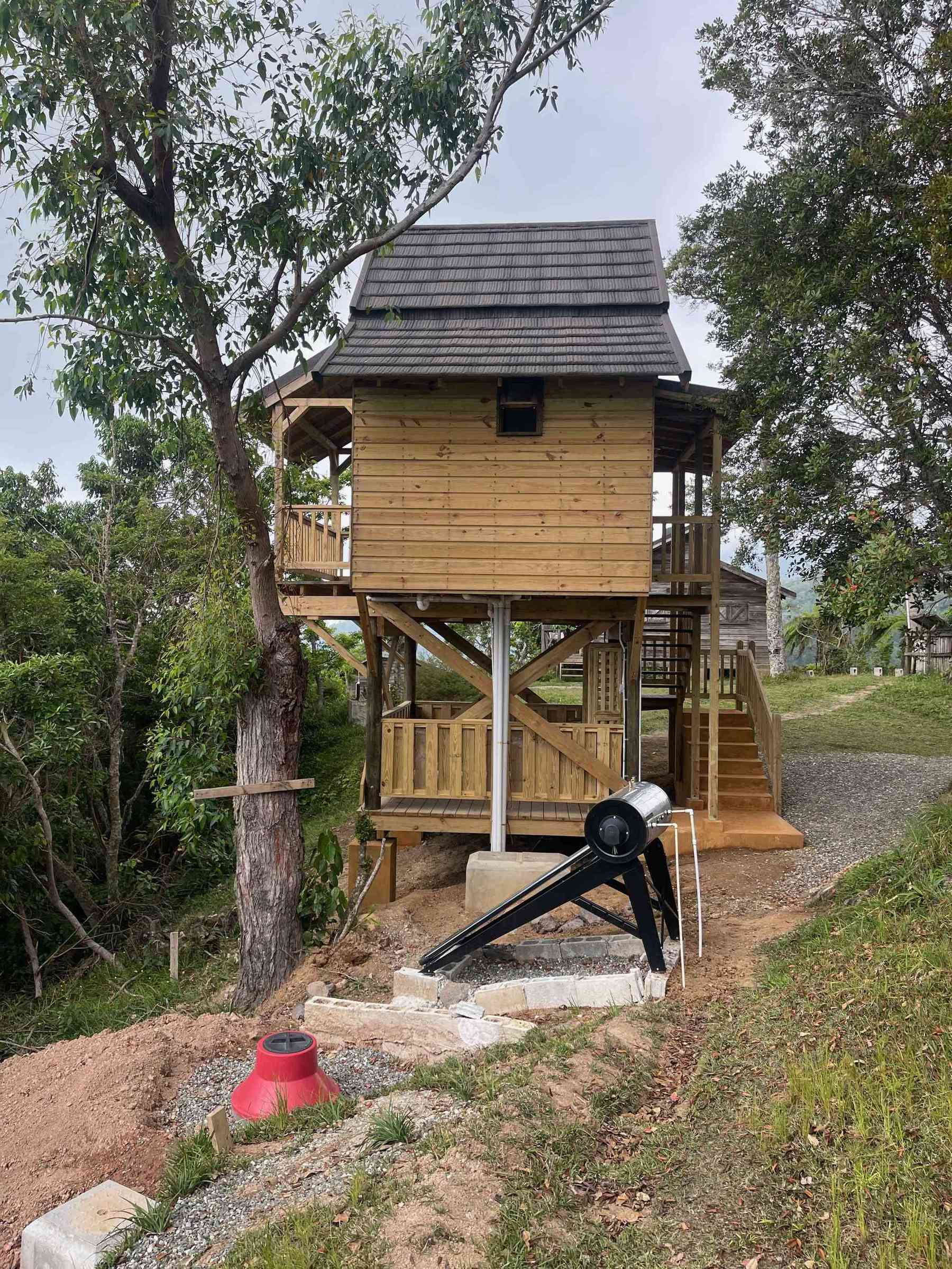 The rear view of an elevated wooden cabin at Holywell Park. A wooden staircase leads up to the second level. In the foreground, there is an installed solar water heater and a red dome-shaped structure near the base of the cabin. The cabin has a dark shingled roof and is framed by trees.