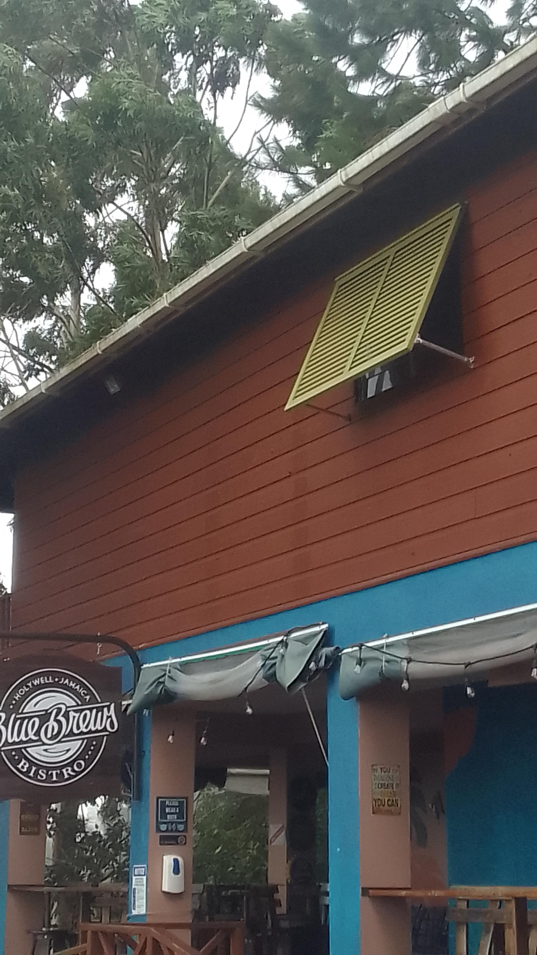 A close-up shot of the upper section of the Blue Brews Bistro building at Holywell Park, Jamaica. The building has reddish-brown wooden siding and a horizontal blue band, with a yellow louvered window awning visible. The sign for "Blue Brews Bistro" is partially visible on the left.