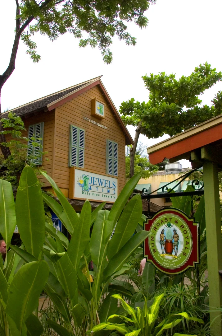 Timber duty-free shop with blue louvered shutters and a “Jewels & Time” sign, rum-roast sign and lush tropical planting at Island Village, Ocho Rios.