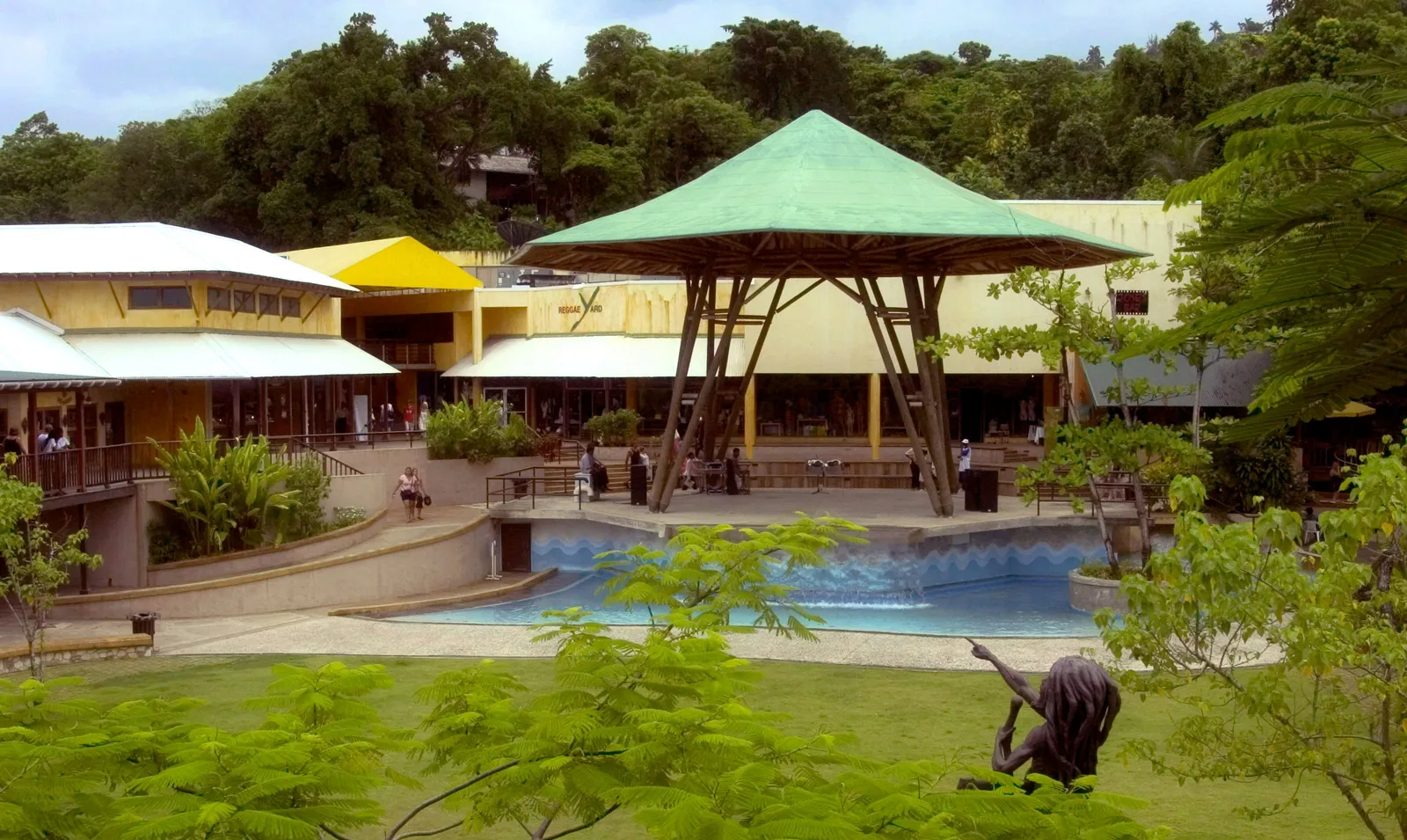 central plaza with tall bamboo canopy over stage, shallow pool and shop-lined promenades set in lush greenery.
