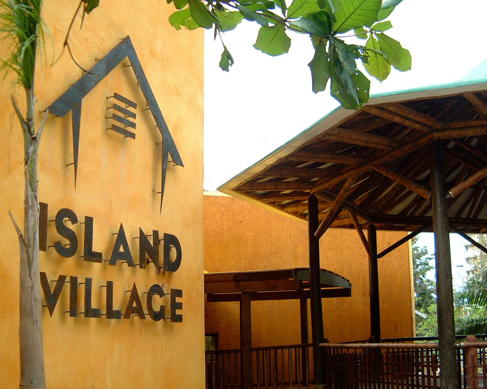 Island Village sign on textured ochre wall beside bamboo-roof walkway and timber railings.