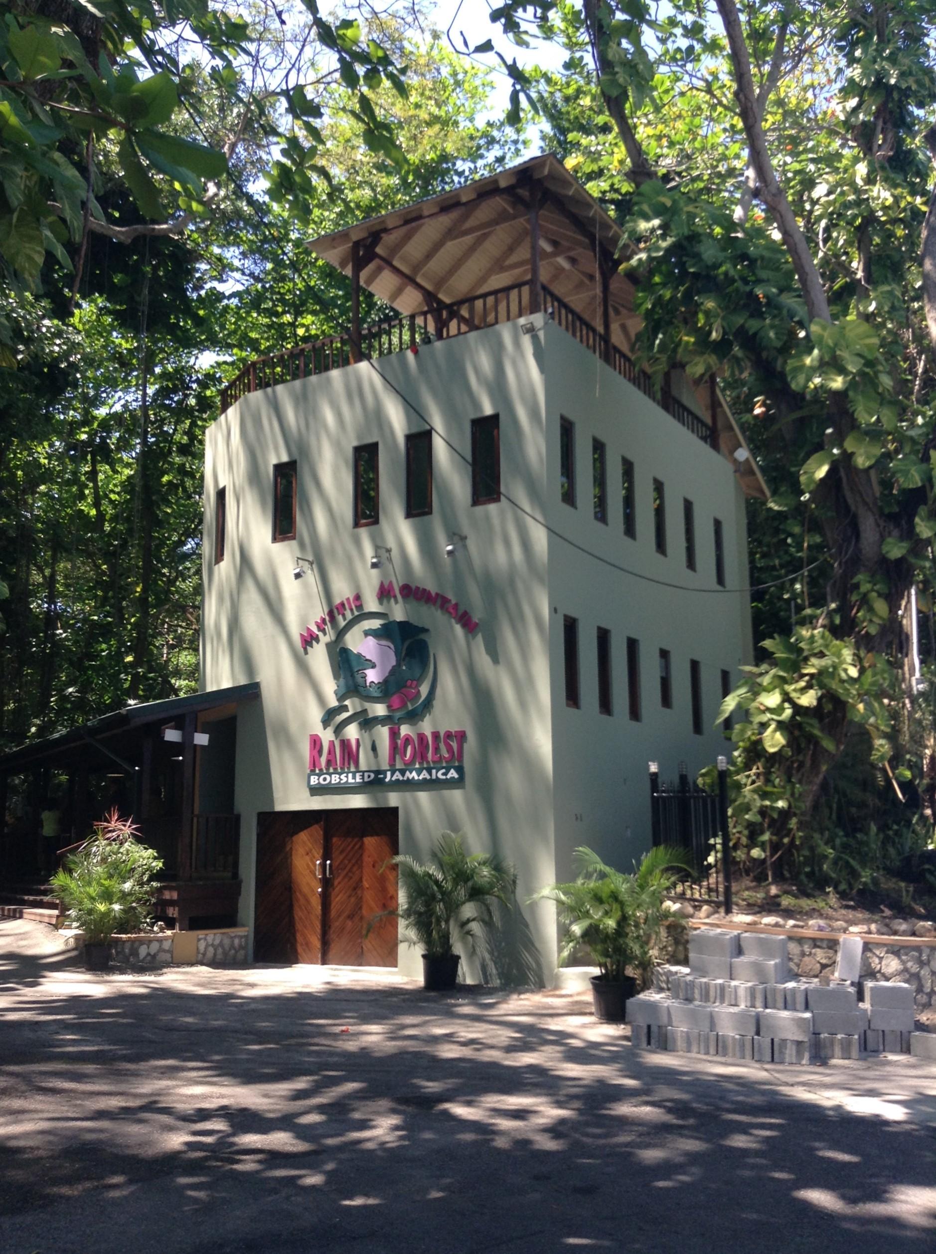 Ground-level view of a light green, four-story building with the "Mystic Mountain Rain Forest Bobsled Jamaica" logo, with a small observation deck on top, surrounded by trees and a paved entrance area.