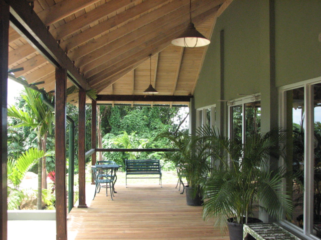 View of an open-air wooden porch with a sloped wooden ceiling, rustic lighting, a small table, a bench, and potted plants, overlooking dense green forest.