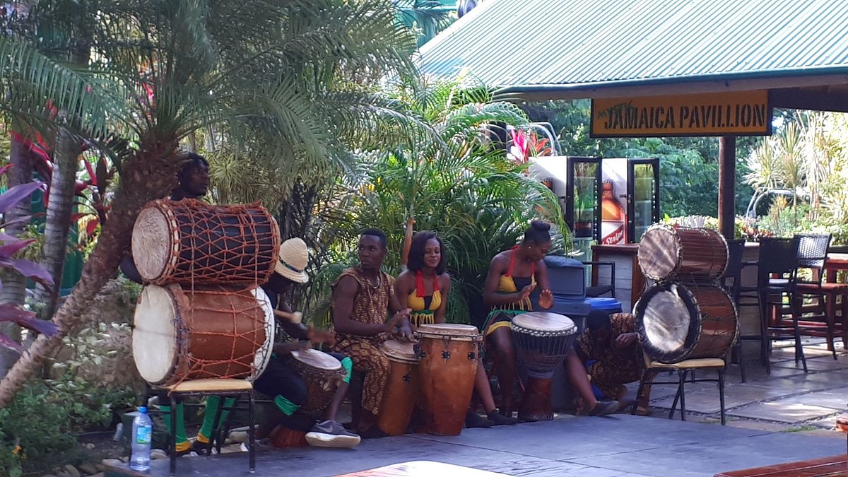 A group of people dressed in traditional-looking attire playing large, vertical drums in an outdoor patio area labeled "JAMAICA PAVILLION" with tropical plants in the background.