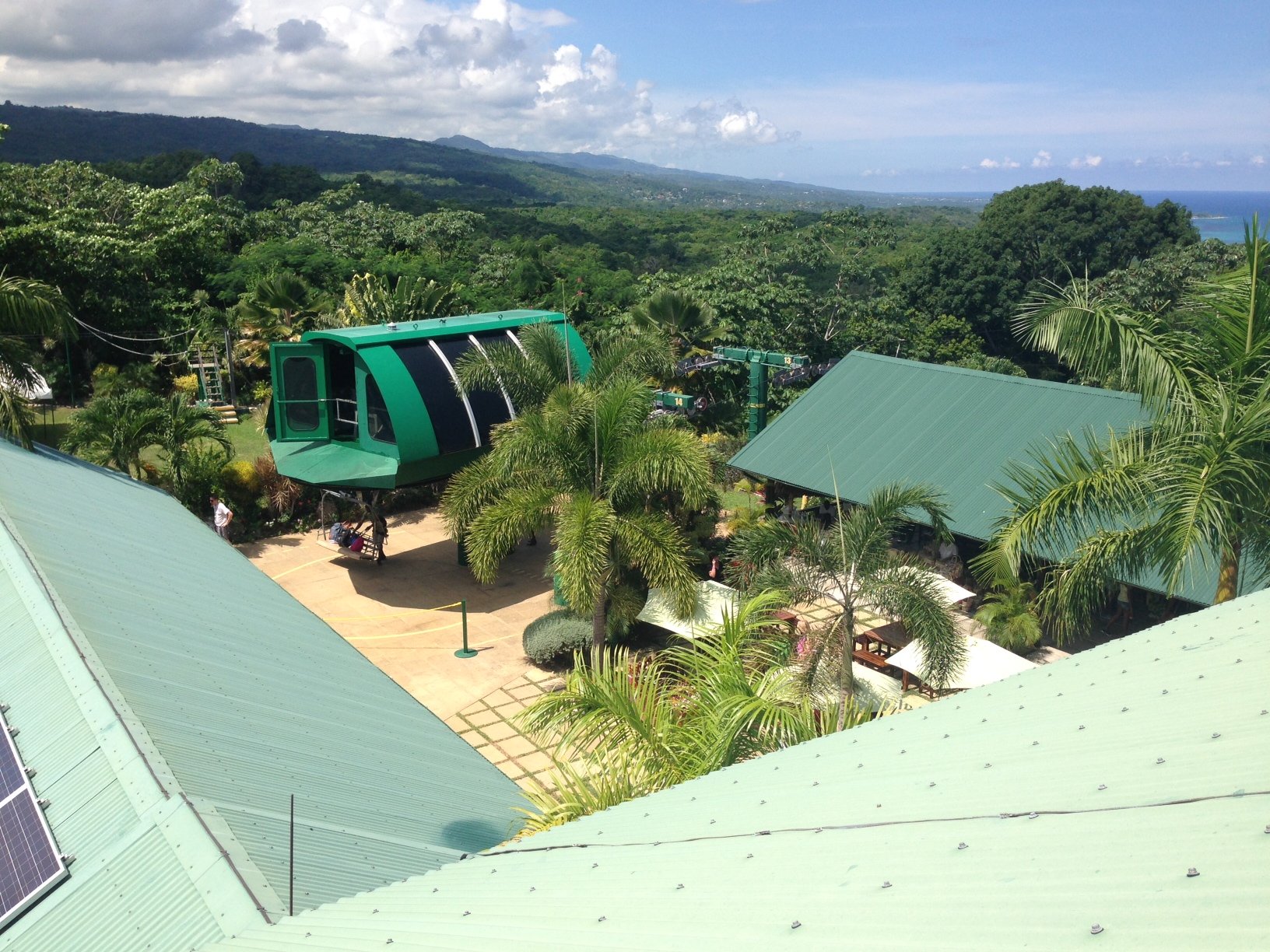 High-angle view over a green-roofed building and a paved courtyard, showing a large green enclosed ski lift gondola station and the lush, hilly terrain of the Jamaican rainforest extending to the blue ocean in the distance under a partly cloudy sky.